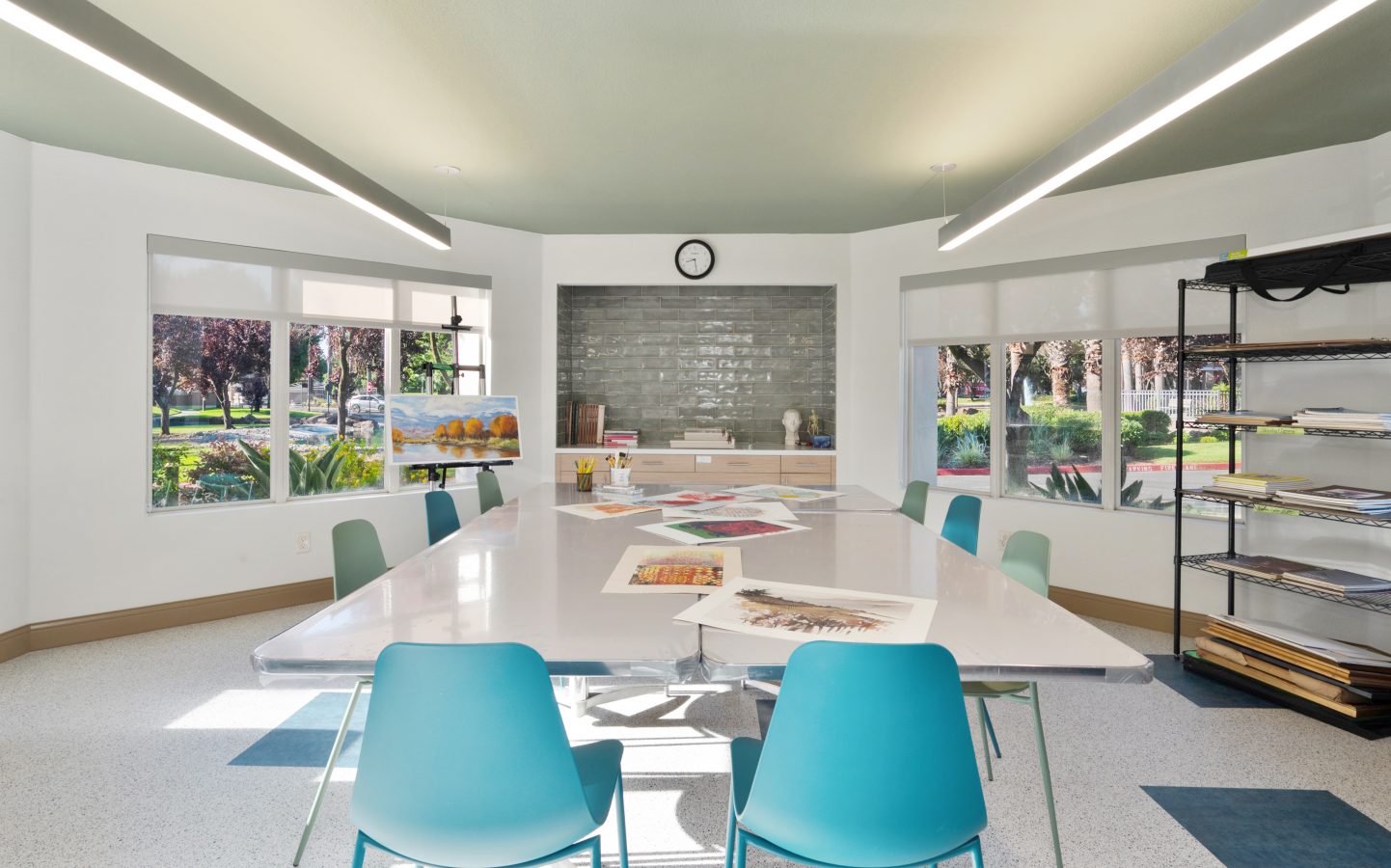 Bright art studio in a Life Plan Community with large windows, teal chairs around a white table covered in colorful artworks, shelves with supplies on the right, and a clock above a tiled backsplash at the back. Greenery visible outside.