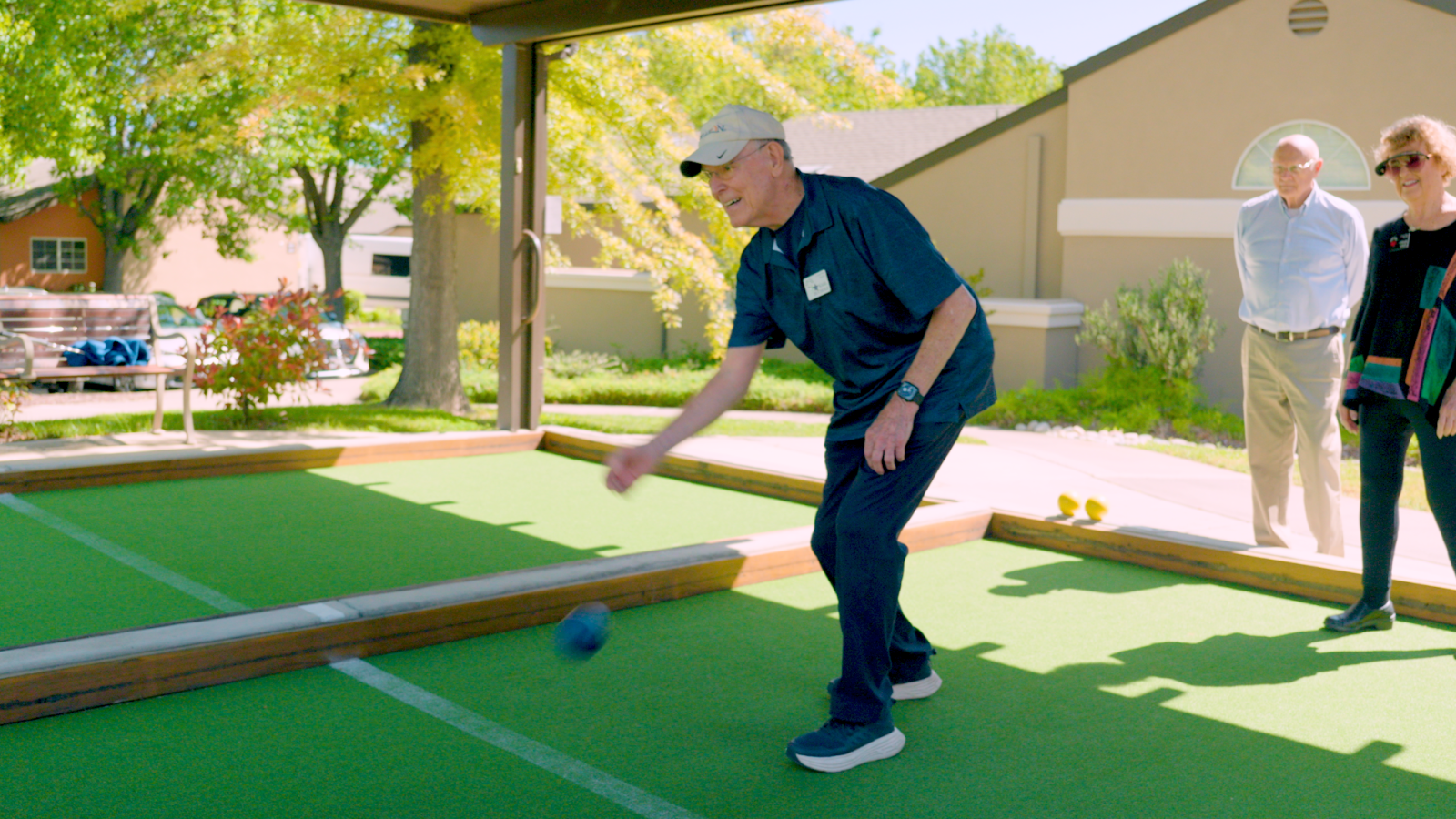 An older man wearing a cap and dark clothes throws a bocce ball on an outdoor court at a vibrant senior living community, while two people stand nearby watching on a sunny day.