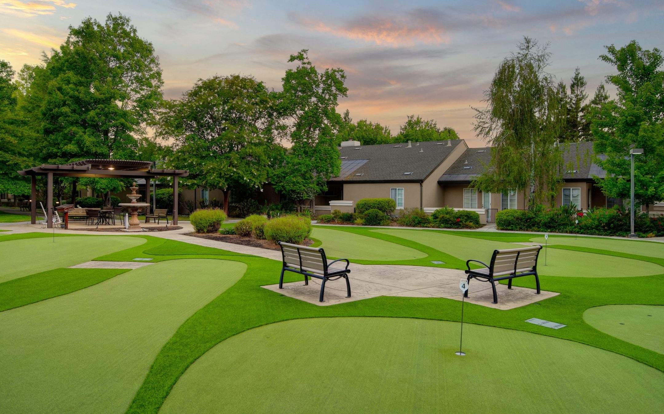 A landscaped putting green area at Eskaton Village Carmichael features benches, a water bottle on one bench, a gazebo with seating, lush trees, and a building in the background at sunset.