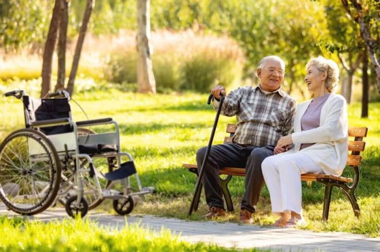 An elderly couple sits on a park bench, smiling and holding hands. The man has a cane, and an empty wheelchair is nearby, reflecting the many living options available for seniors to enjoy sunny, green days together.