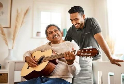 An older man smiles while playing an acoustic guitar, seated on a bench, as a younger man stands beside him, leaning on the backrest and smiling warmly—showcasing joyful moments shared while exploring living options in a bright, cozy room.