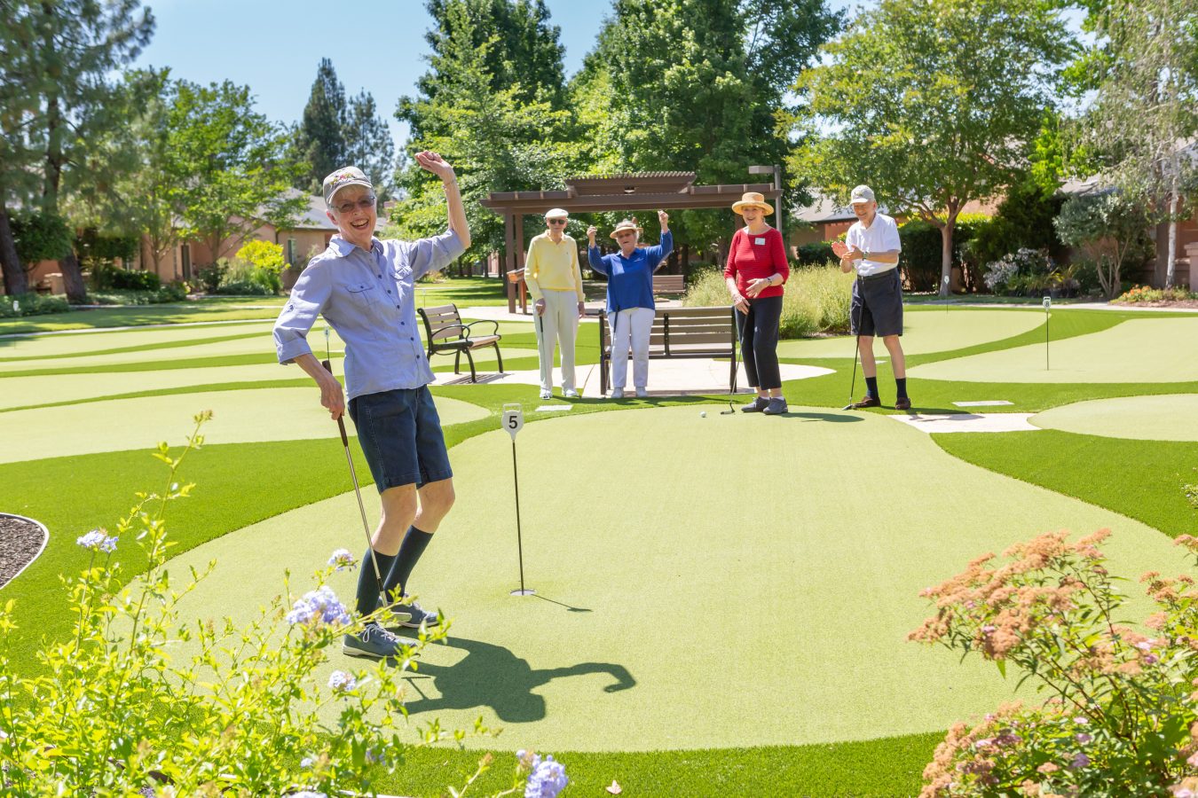 A group of older adults enjoy a sunny day playing mini golf on a well-maintained putting green, with one person celebrating and others watching or preparing to putt. Trees and flowers surround the cheerful scene.