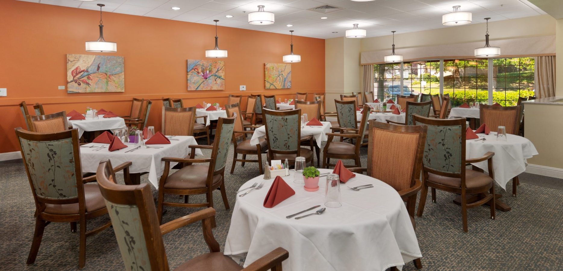 A dining room with round tables covered in white tablecloths, set for a memorable culinary experience with red napkins, glasses, and silverware. Wooden chairs surround the tables, while orange walls and colorful paintings create a warm, inviting atmosphere.