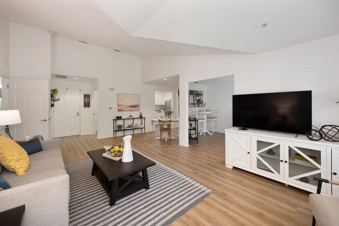 A spacious living room with light wood floors, a gray couch with yellow pillows, and a black coffee table offers comfort for short-term stays, featuring a large flat-screen TV on a white console and an open view into the dining area and kitchen.