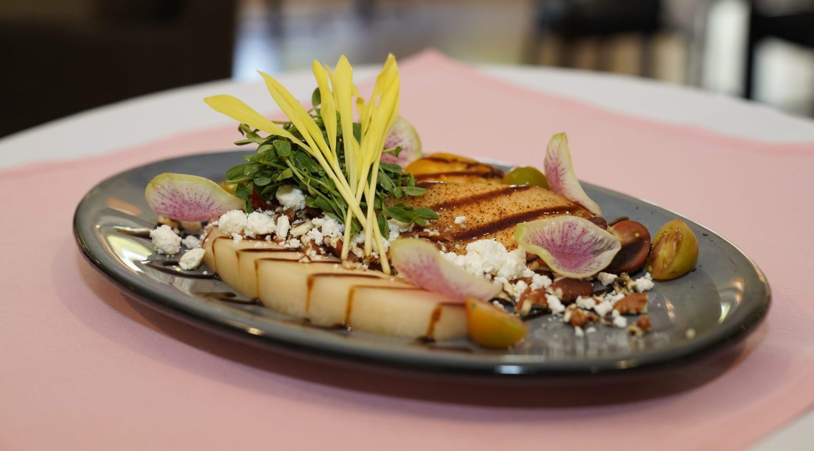 A beautifully plated gourmet dish features sliced melon, yellow flower petals, greens, cheese crumbles, and thinly sliced radishes on a gray plate, set on a pink tablecloth.