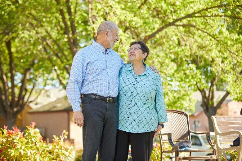 An elderly couple stands outdoors, smiling at each other with arms around one another, surrounded by greenery and sunlight—a beautiful moment reflecting the warmth of Resident Culture. Empty chairs are visible nearby.