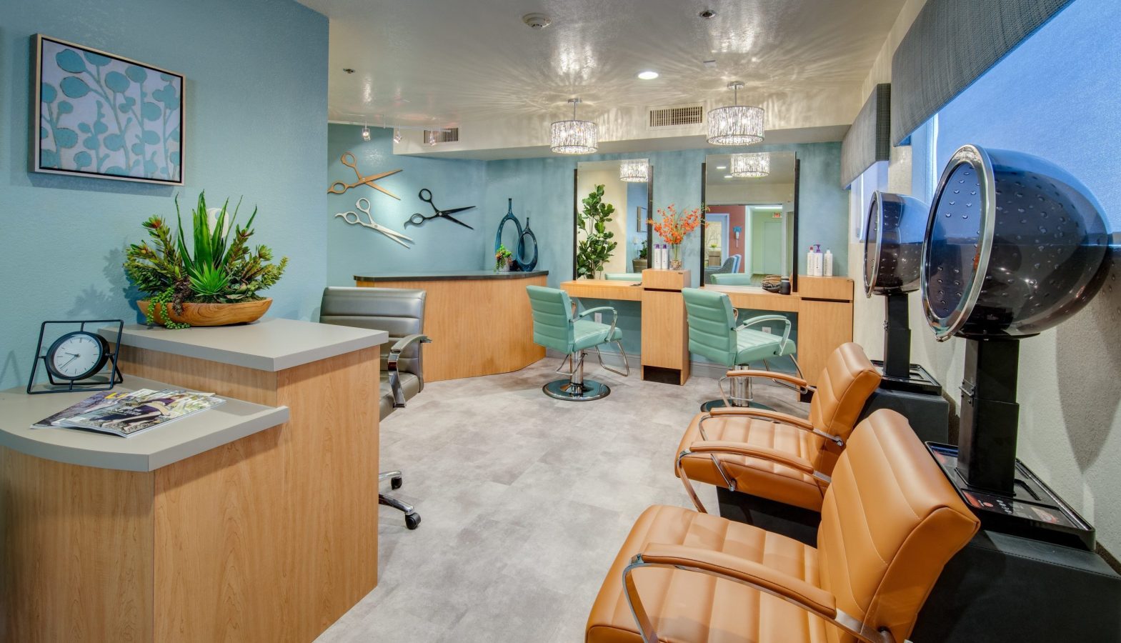 Modern hair salon with brown and teal decor, two styling stations with mirrors and chairs, two hooded dryers, plants, wall art, and a reception desk with magazines and a clock. Large windows let in natural light.