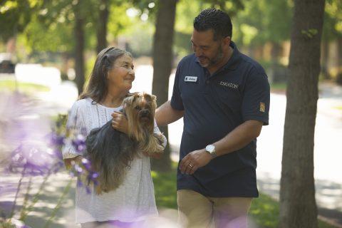 An older woman holding a small dog stands outdoors, smiling at a man in a navy polo shirt with a name tag. They are surrounded by trees and flowers on a sunny day.