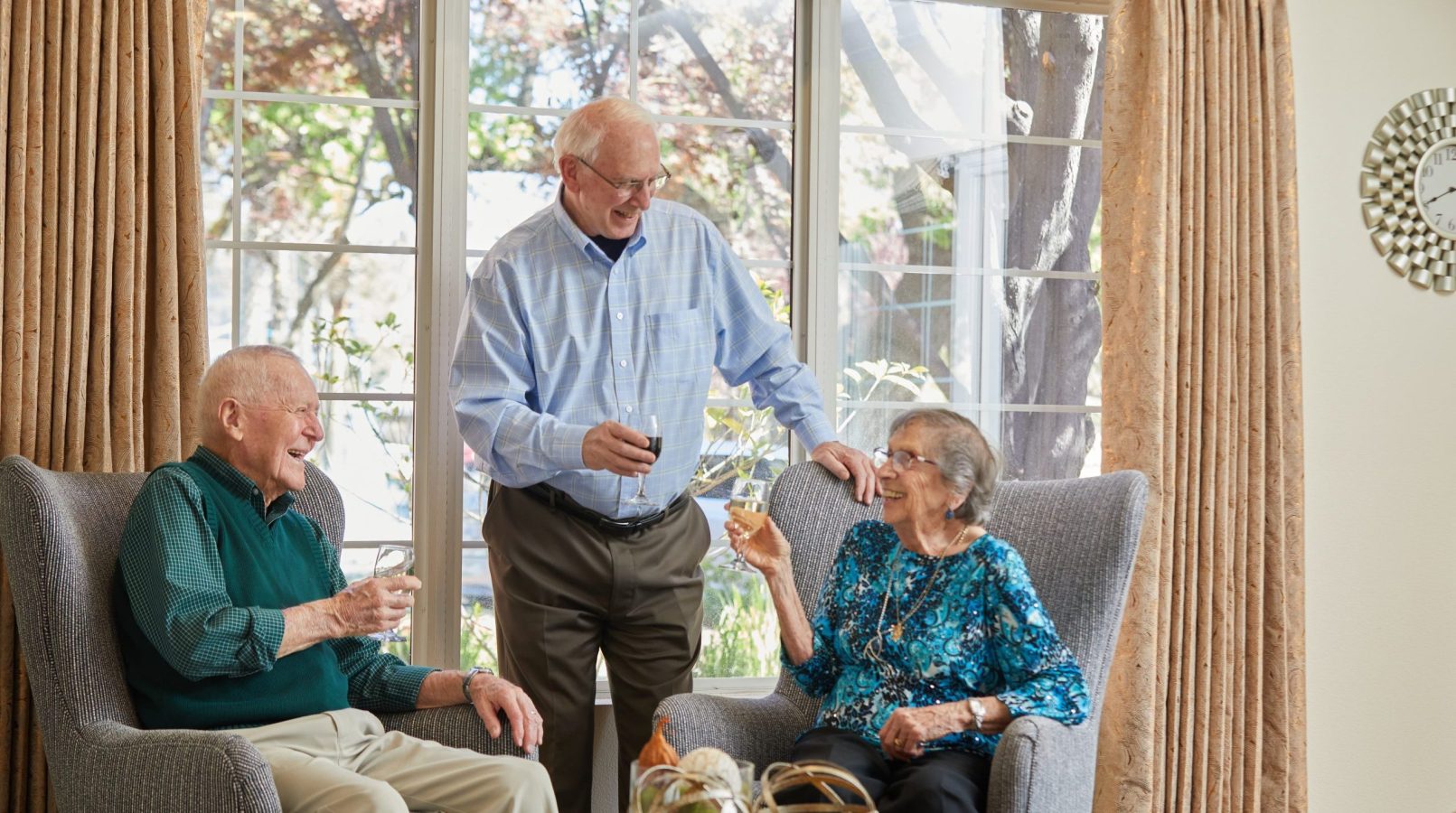 Three smiling seniors enjoy drinks together in a bright living room with large windows and beige curtains. Two sit in armchairs while one stands, all engaged in cheerful conversation, reflecting the vibrant resident culture.
