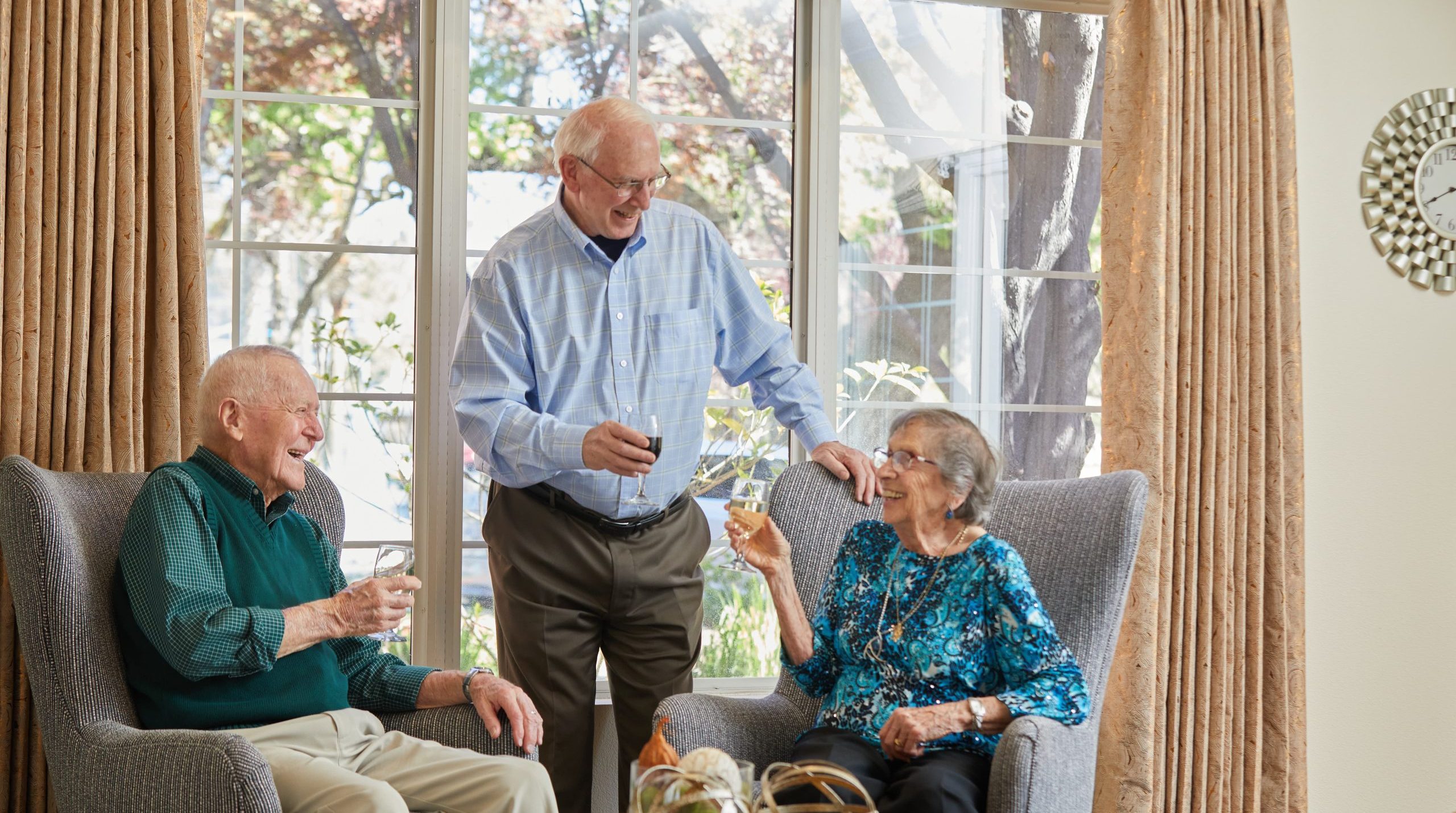 Three smiling seniors enjoy drinks together in a bright living room with large windows and beige curtains. Two sit in armchairs while one stands, all engaged in cheerful conversation, reflecting the vibrant resident culture.