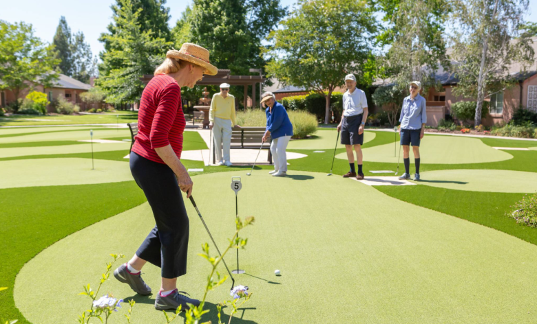 Five older adults are playing mini-golf on a bright, sunny day. One woman in a red shirt and straw hat prepares to putt while the others watch. The putting green is surrounded by trees and landscaped gardens.