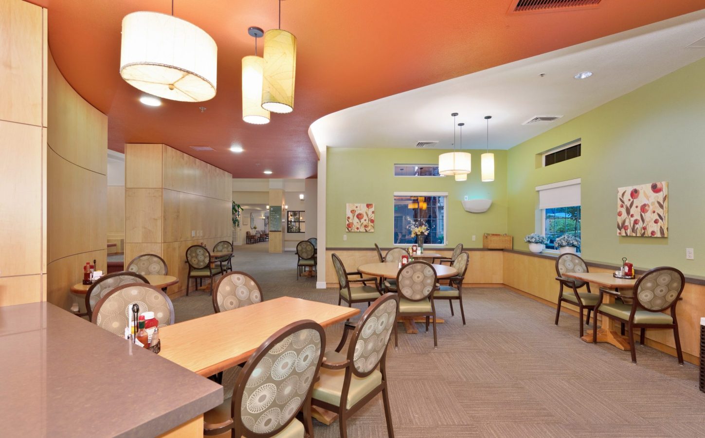 A brightly lit dining area with round and square tables, cushioned chairs, modern pendant lights, and green and orange walls decorated with floral art sets the stage for a memorable culinary experience. Large windows provide an outdoor view.