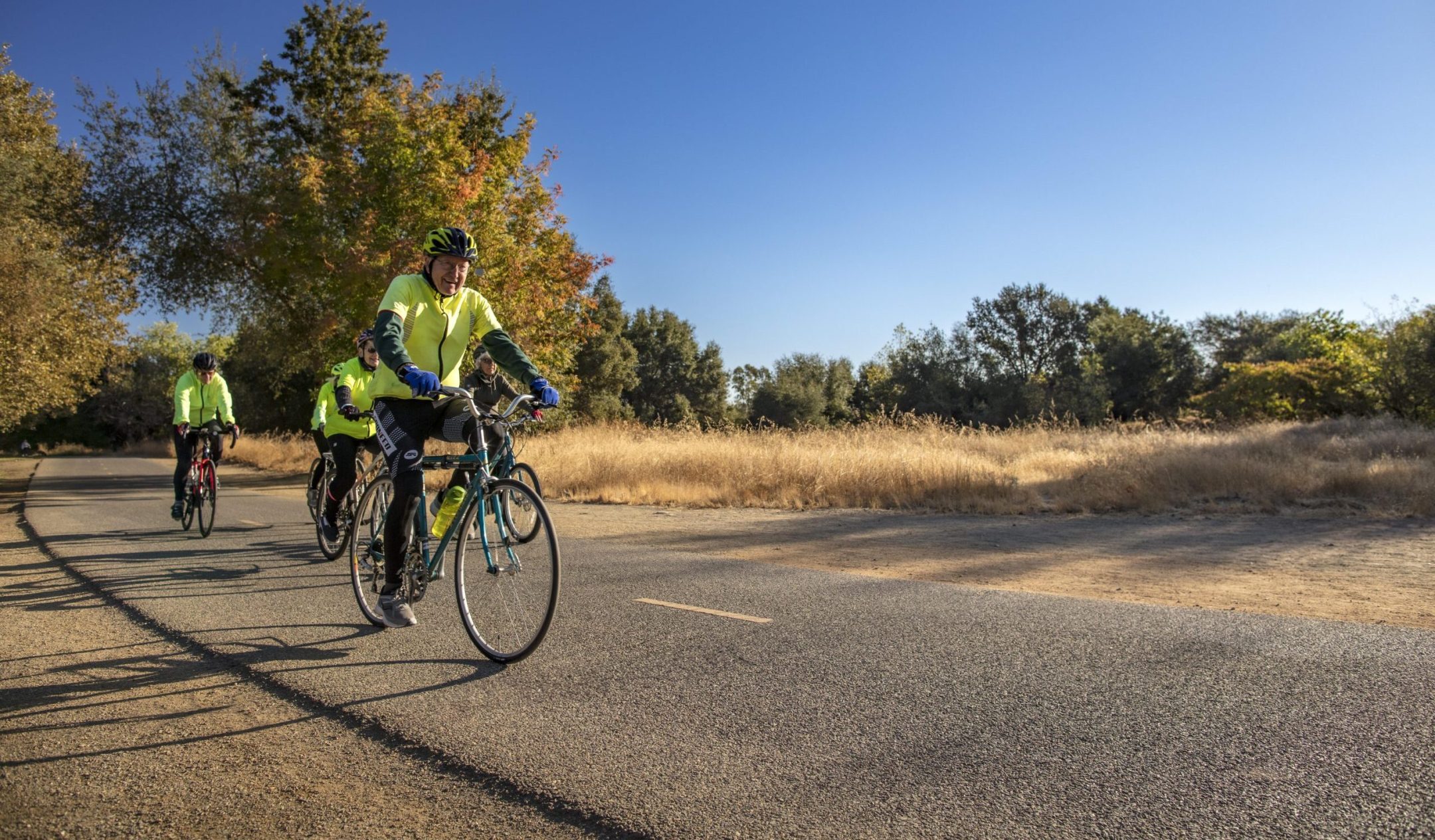 Three cyclists in bright yellow jackets and helmets ride along a paved path through a park, embracing the resident culture of outdoor activity amid dry grass, autumn trees, and a clear blue sky.