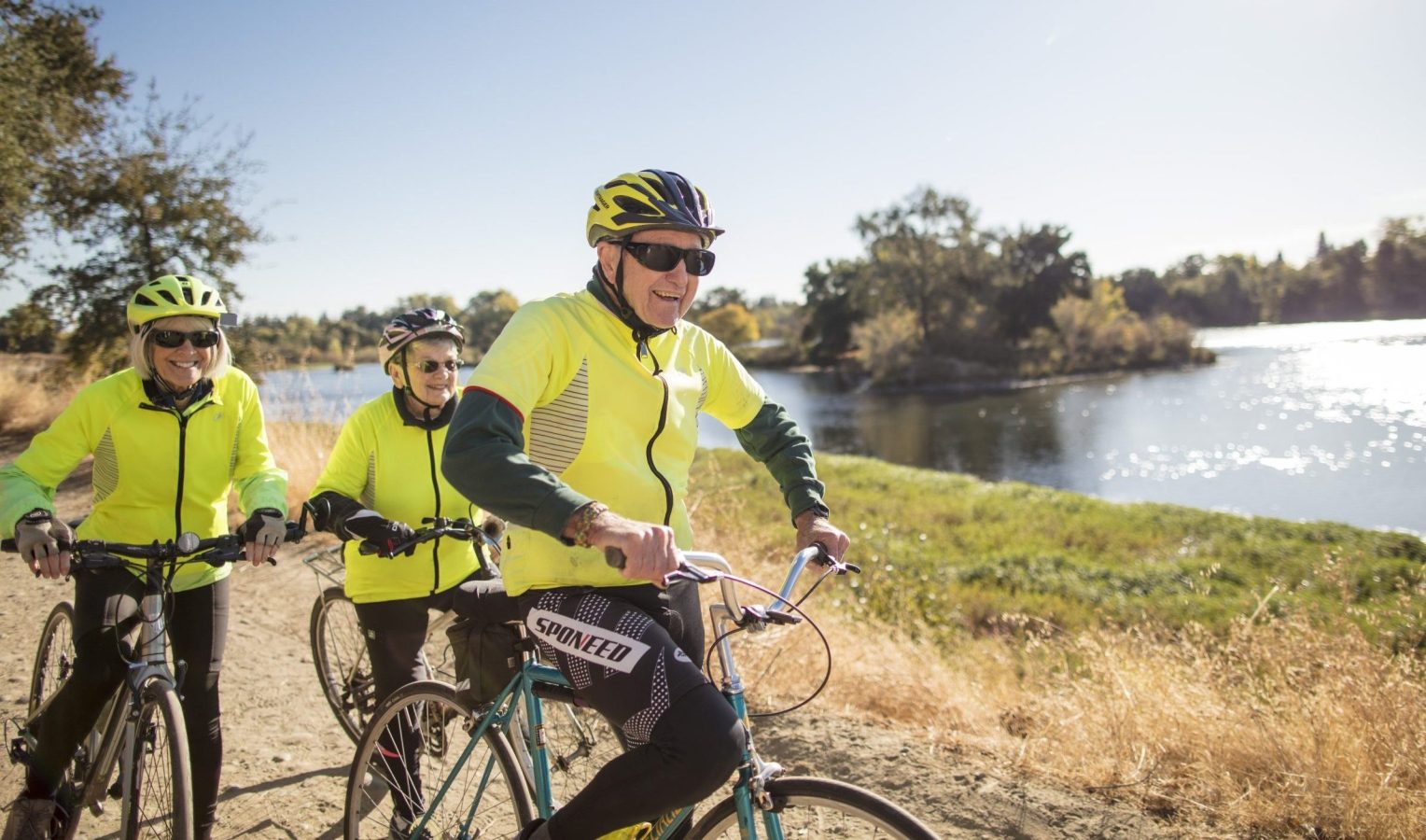 Three older adults wearing bright yellow jackets and helmets ride bicycles along a sunlit riverside trail, surrounded by grass and trees on a clear day.