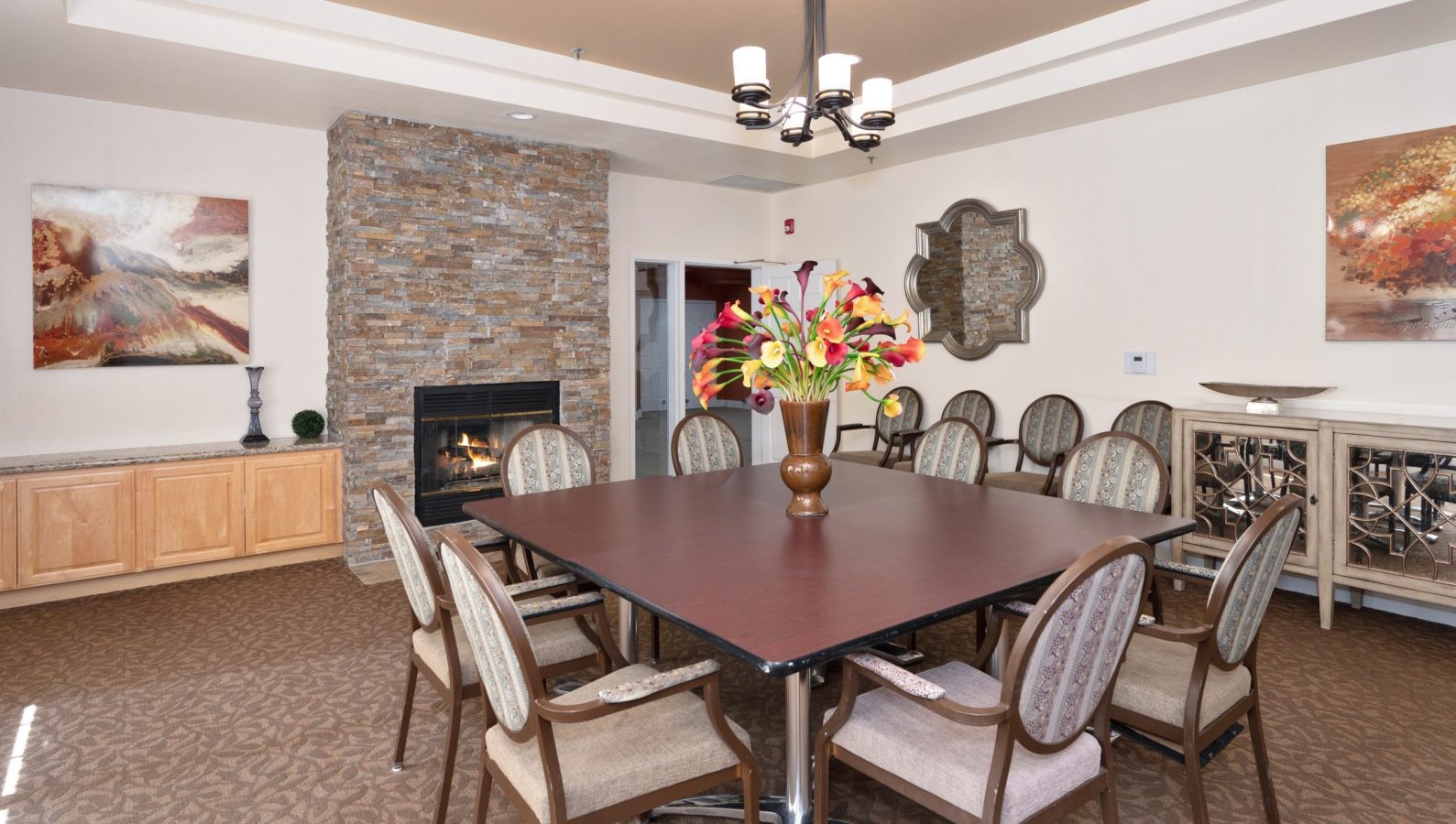 A dining room designed for a memorable culinary experience, featuring a square table with ten upholstered chairs, a vibrant vase of flowers at the center, a stone fireplace, decorative art on the walls, and a chandelier overhead.