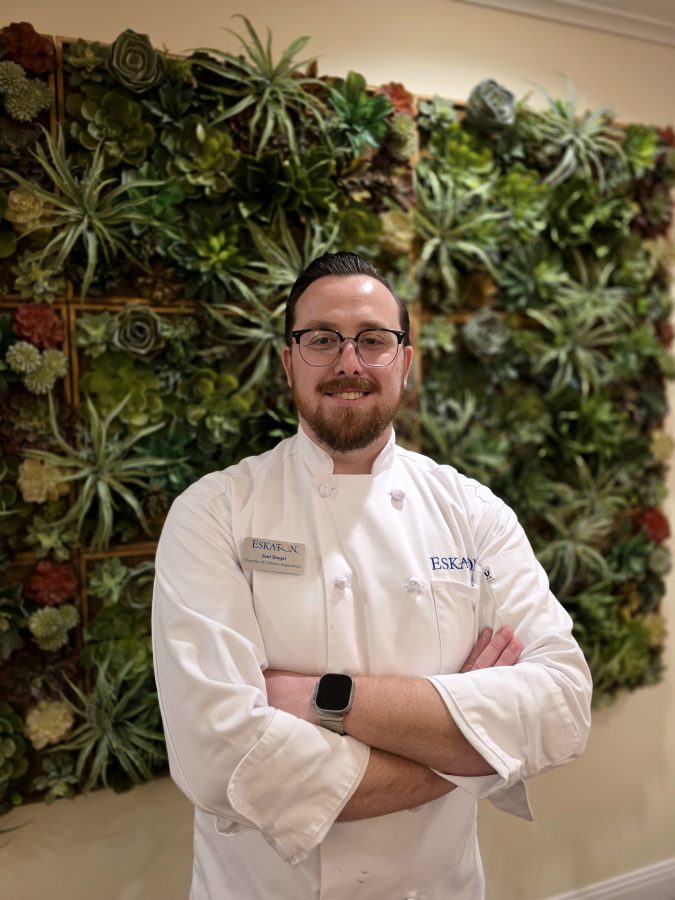 A chef wearing a white uniform and glasses stands with arms crossed in front of a wall decorated with green and red succulents, showcasing the vibrant atmosphere of Eskaton Village Carmichael dining.