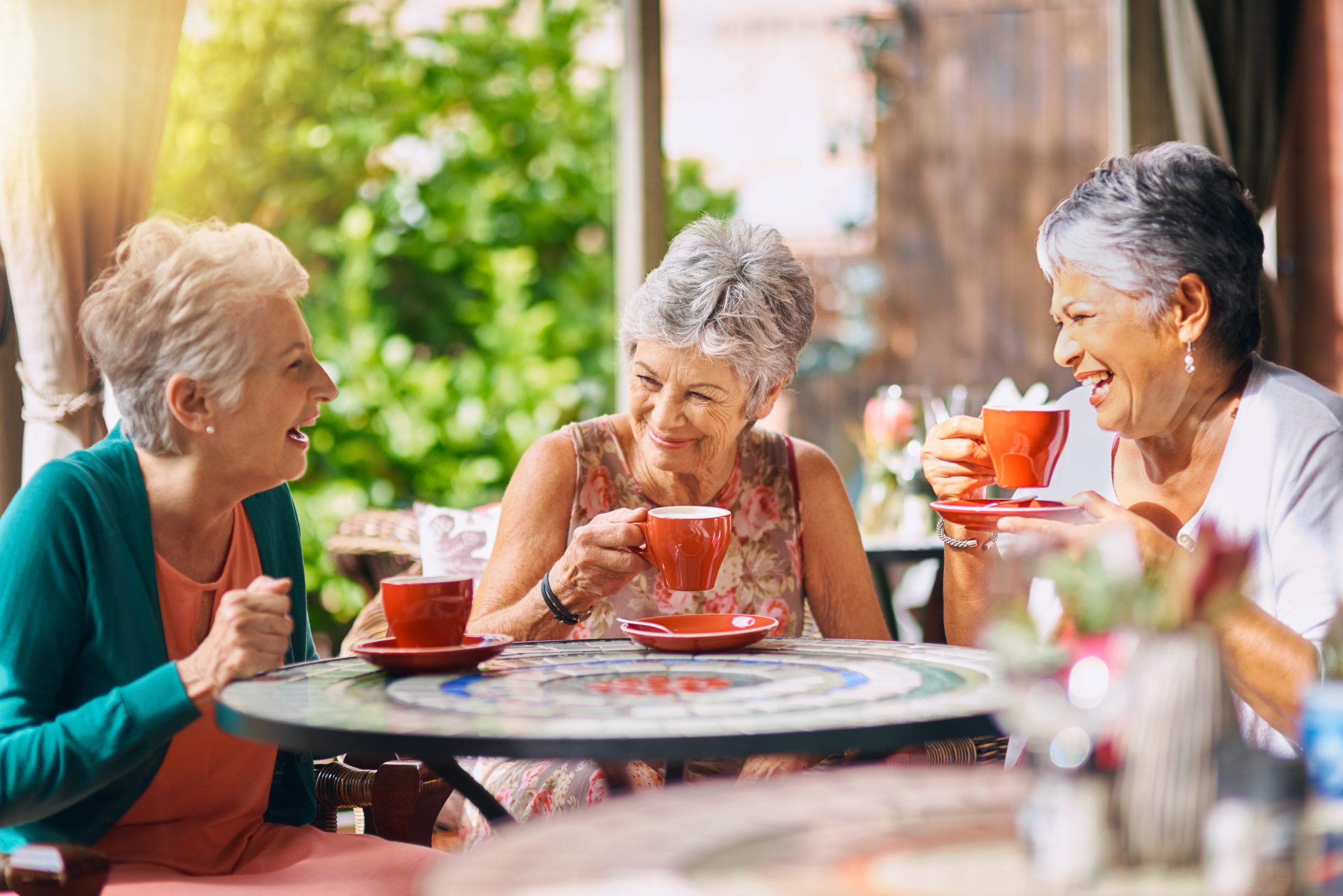 Three elderly women sit at a round outdoor table, smiling and laughing together while drinking from bright orange cups, enjoying a sunny day surrounded by greenery.