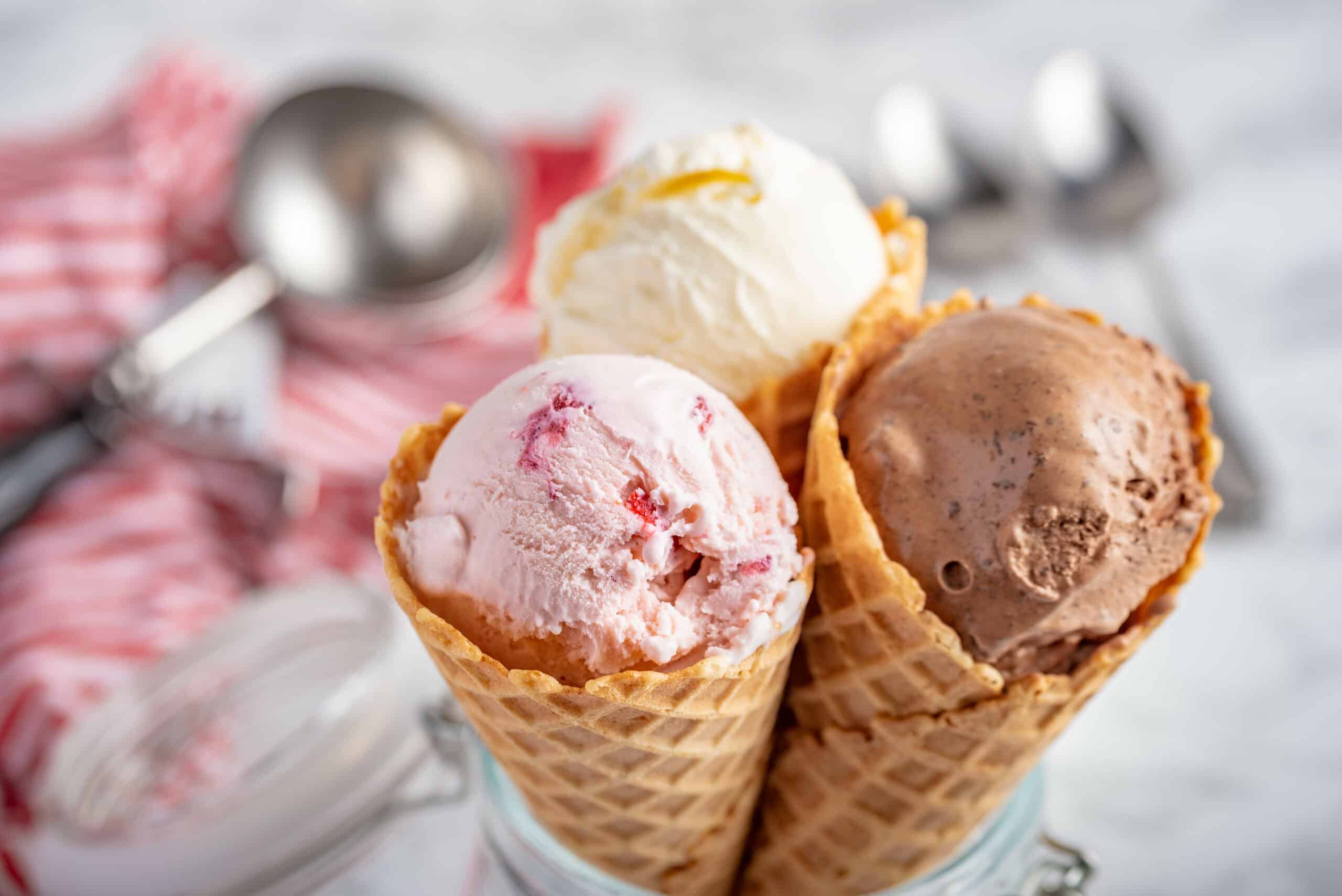 Three waffle cones filled with vanilla, strawberry, and chocolate ice cream are displayed in a glass jar—perfect for an Ice Cream Social. The background features metal scoops and a red-striped cloth for a festive touch.