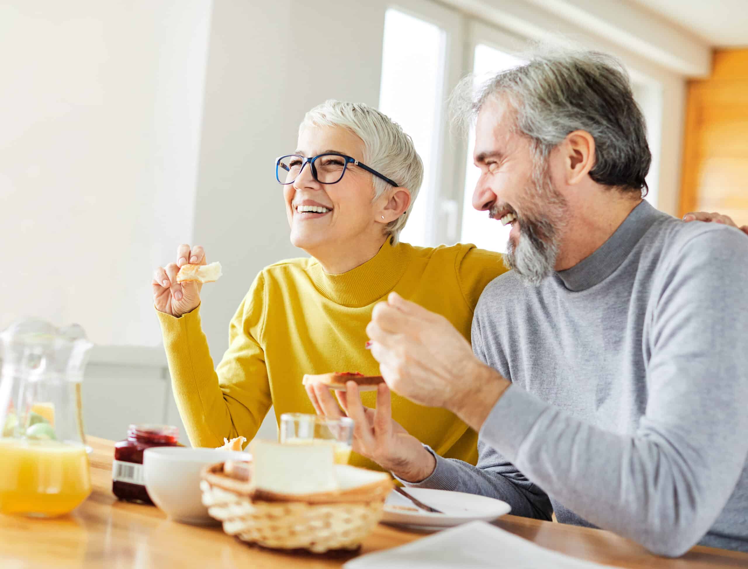 An older woman and man sit together at a table, smiling and enjoying breakfast as they discuss their life plan. The woman wears glasses and a yellow sweater; the man wears a gray sweater. Baskets of bread and jars of jam are on the table.