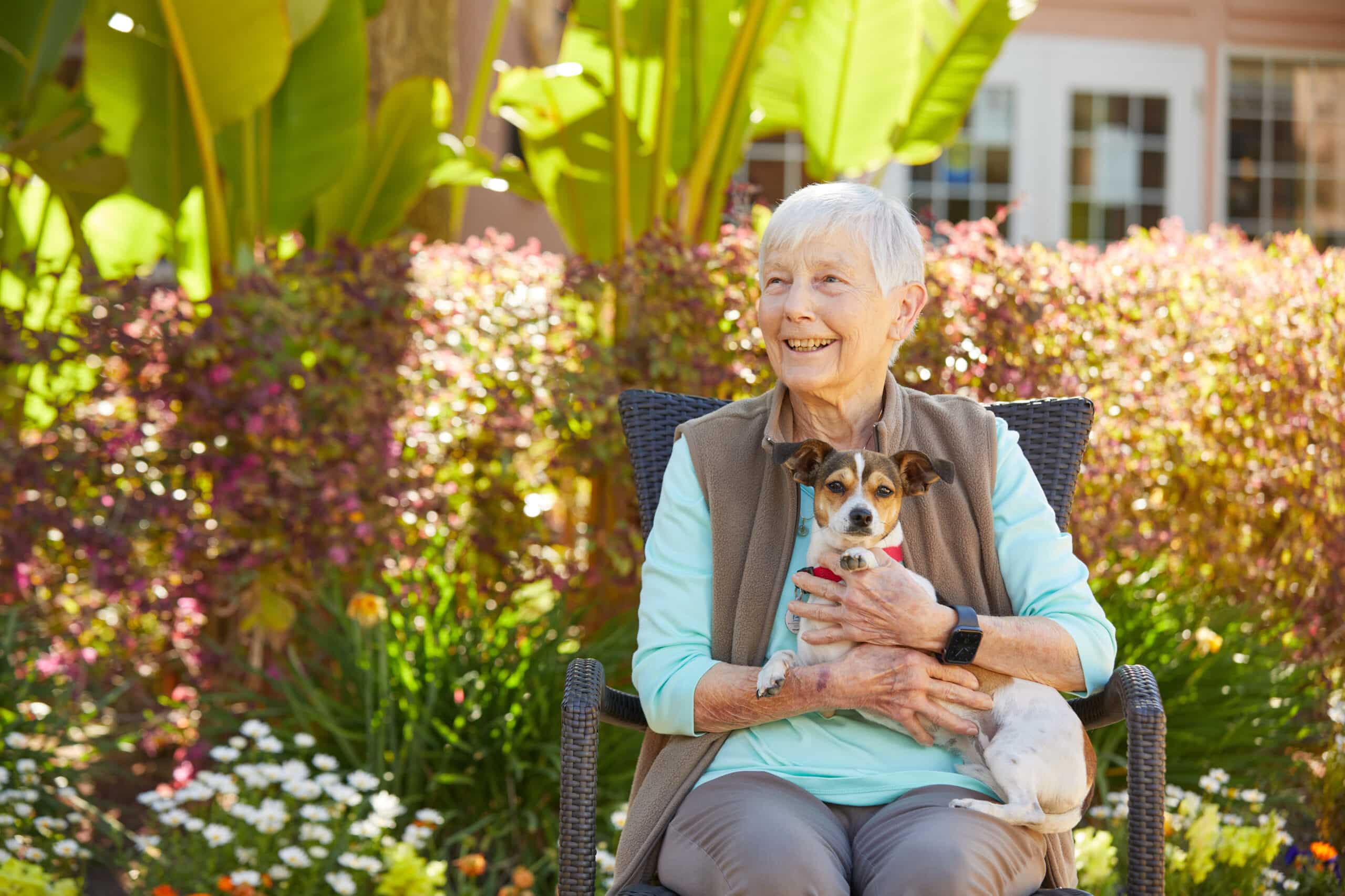 Smiling older woman sits outdoors in a chair, holding a small dog on her lap. She is surrounded by colorful plants and flowers, with sunlight illuminating the garden scene—a heartwarming moment captured during the Community Spotlight Event.