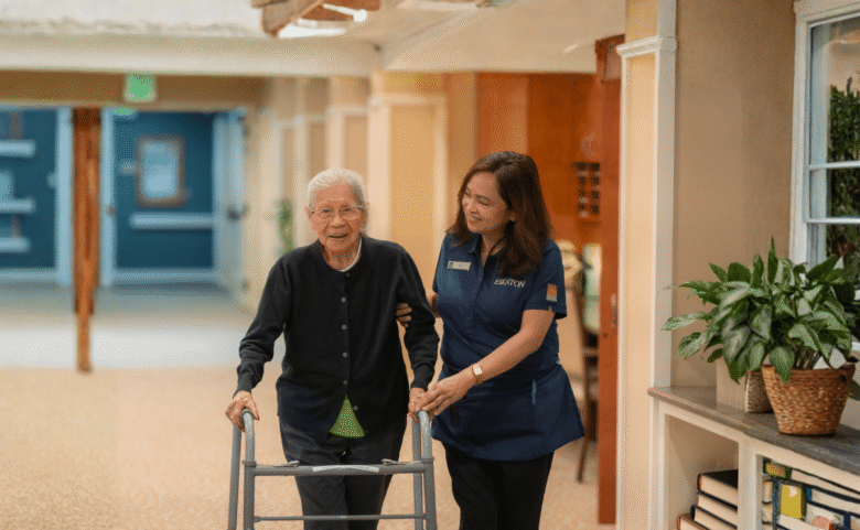 An elderly woman using a walker is assisted by a caregiver in a hallway with warm lighting, plants, and bookshelves.