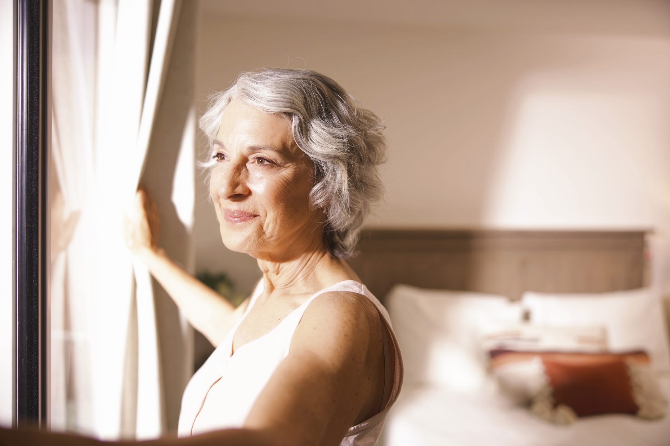 An older woman with short gray hair stands by a window, gently holding open white curtains. Sunlight shines on her face, and there is a neatly made bed in the softly lit background.