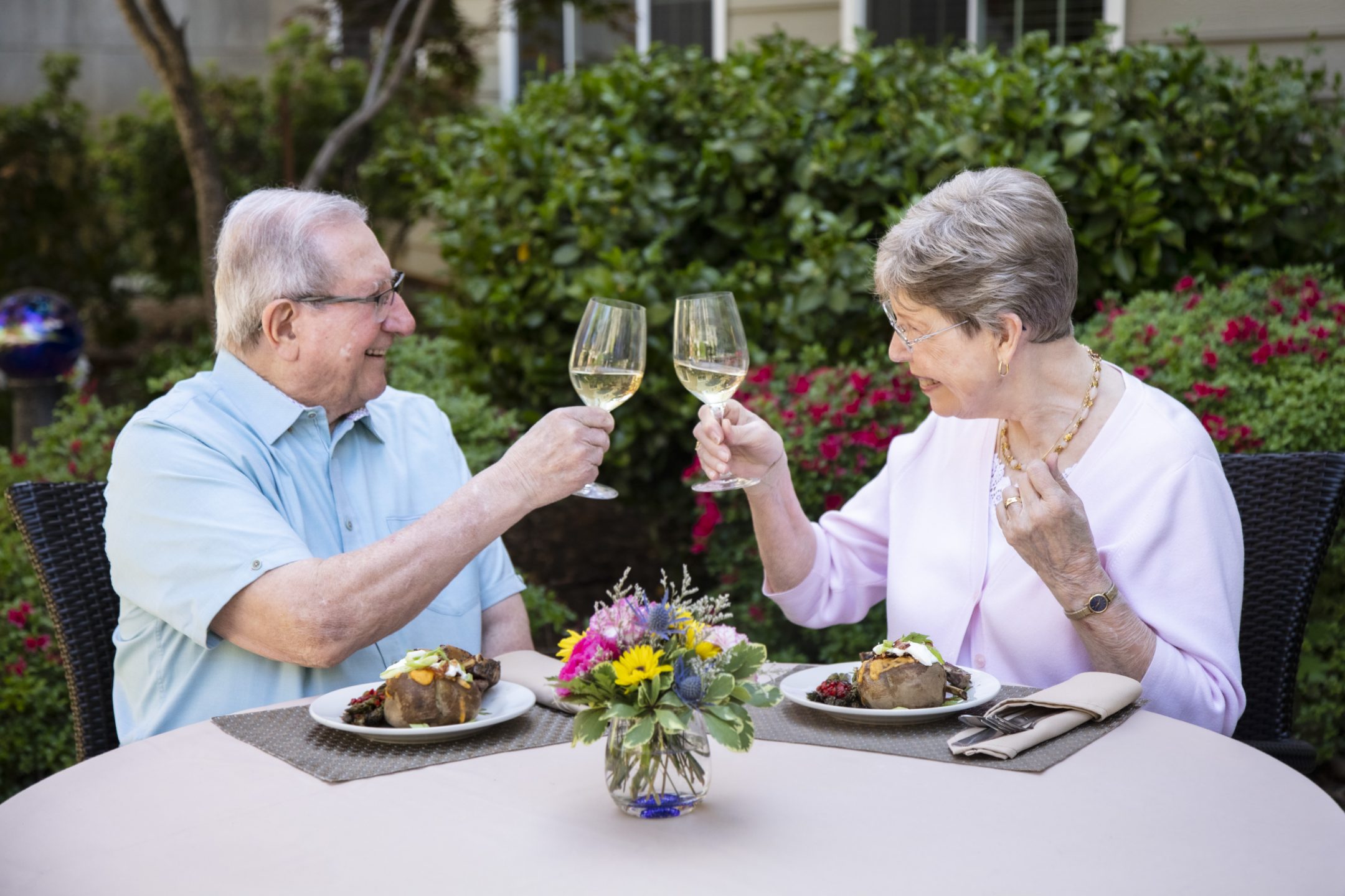 An elderly man and woman sit at an outdoor table, smiling and clinking wine glasses over plates of food. A small bouquet of flowers sits in the center of the table, with greenery and flowers in the background.
