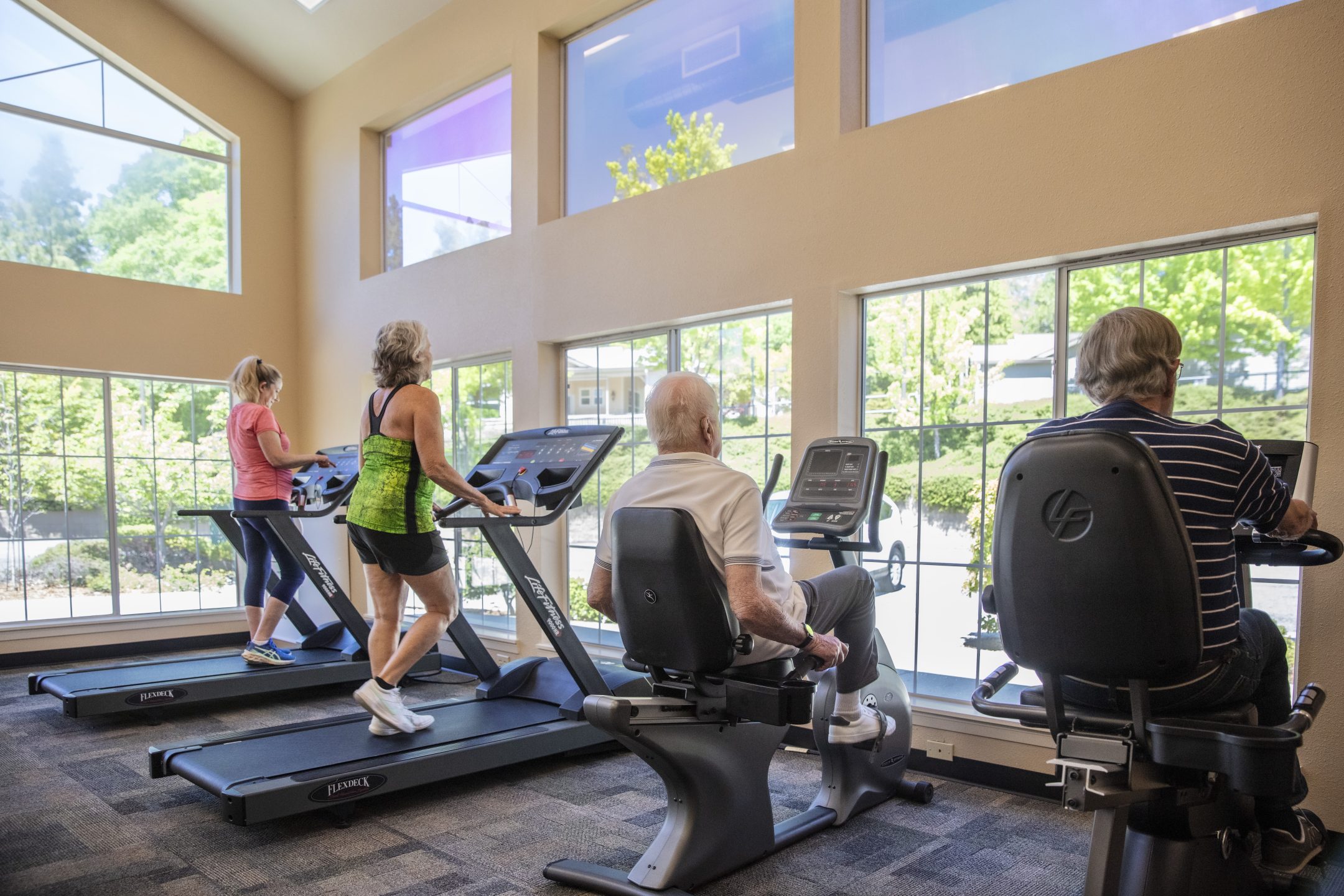 Four older adults exercise in a bright gym with large windows at Eskaton Lifestyle Grass Valley. Two walk on treadmills while two others use stationary bikes, enjoying views of trees and greenery outside.