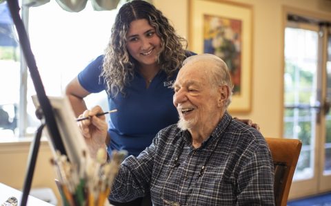 An elderly man smiles while painting on a canvas, as a woman with curly hair and a blue shirt stands beside him, also smiling, in a bright room with artwork on the walls, showcasing creative living options for seniors.