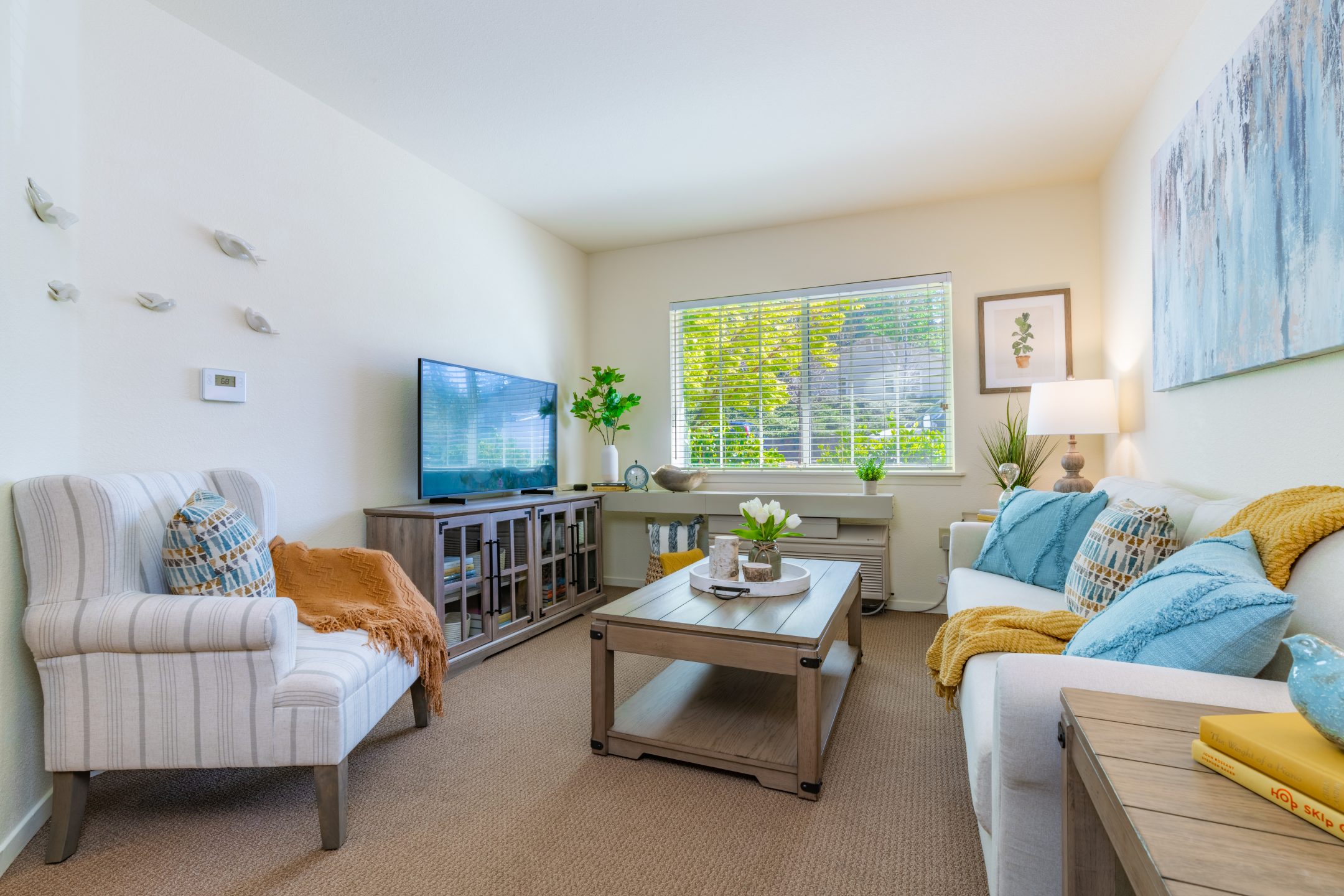 Bright living room with neutral walls, a large window, cozy armchair, sofa with blue and yellow pillows—ideal for short-term stays. Features a wooden coffee table, TV on a console, plants, wall art, and decorative items.
