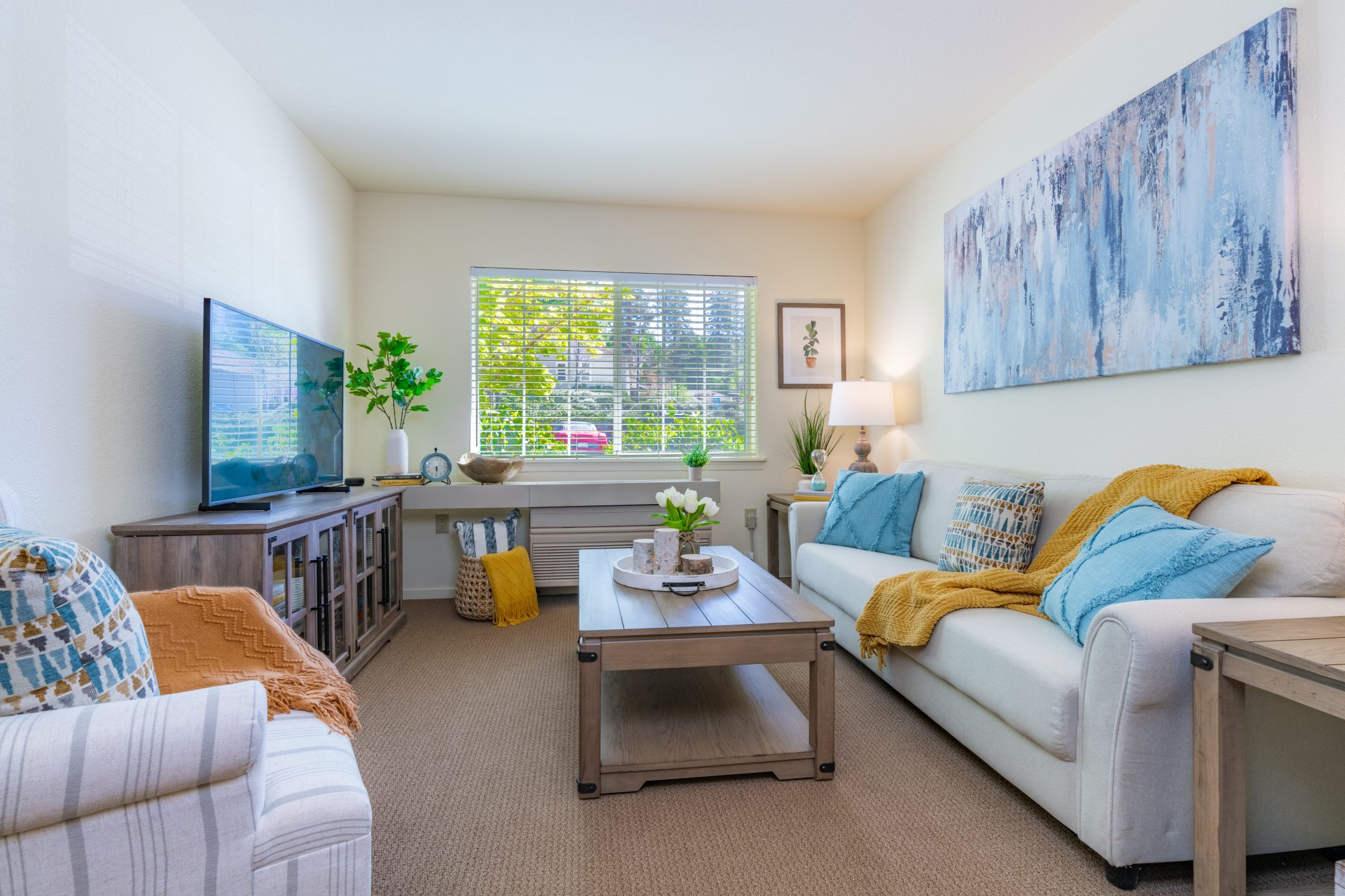 A bright living room at Eskaton Village Grass Valley features a white sofa with blue and yellow cushions, a wooden coffee table, TV, armchair, and large window with blinds. Abstract art hangs above the sofa, while plants decorate the space.