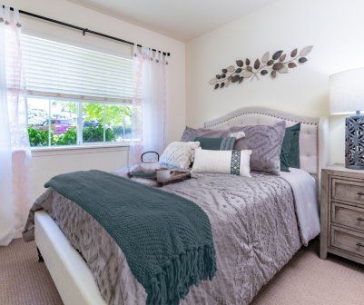 Cozy bedroom at Eskaton Village Placerville with a neatly made bed in gray and teal bedding, a tray with a cup and teapot, natural light from white blinds, a lamp on the nightstand, and decorative wall leaves—perfect for short-term stays.
