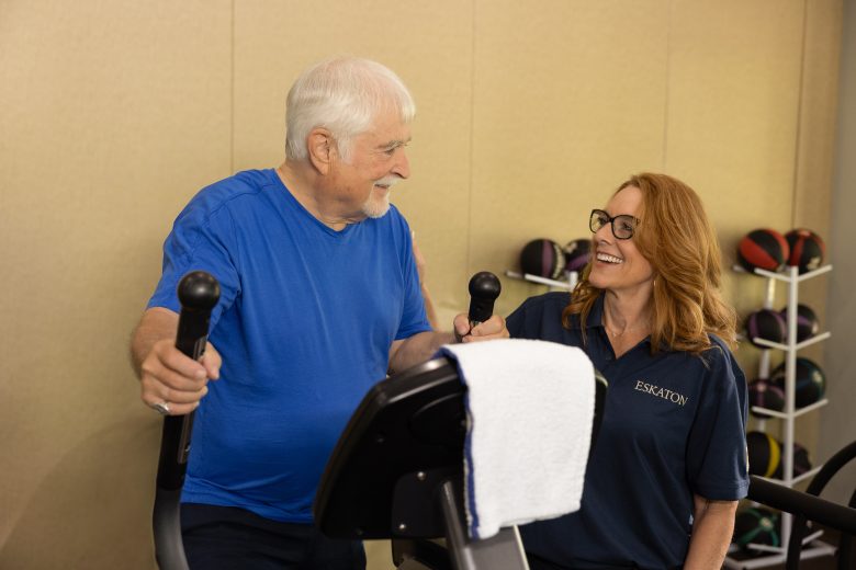 An older man enjoys the Eskaton Village Grass Valley resident experience, smiling and chatting with a woman in glasses and an ESKATON shirt as he uses an exercise machine. Several medicine balls are stacked on a rack in the background.