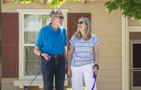 An older man and woman smile at each other while walking two dogs on leashes in front of a house with a tree and landscaping.