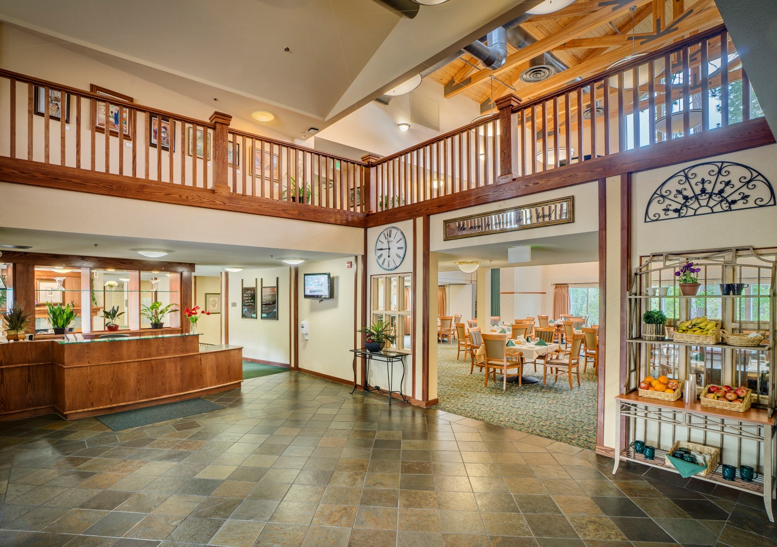 Spacious lobby with tile flooring, wooden railings, a clock on the wall, reception desk, potted plants, metal fruit rack, and an open view to a dining area with tables and chairs at Eskaton Village Grass Valley memory care.