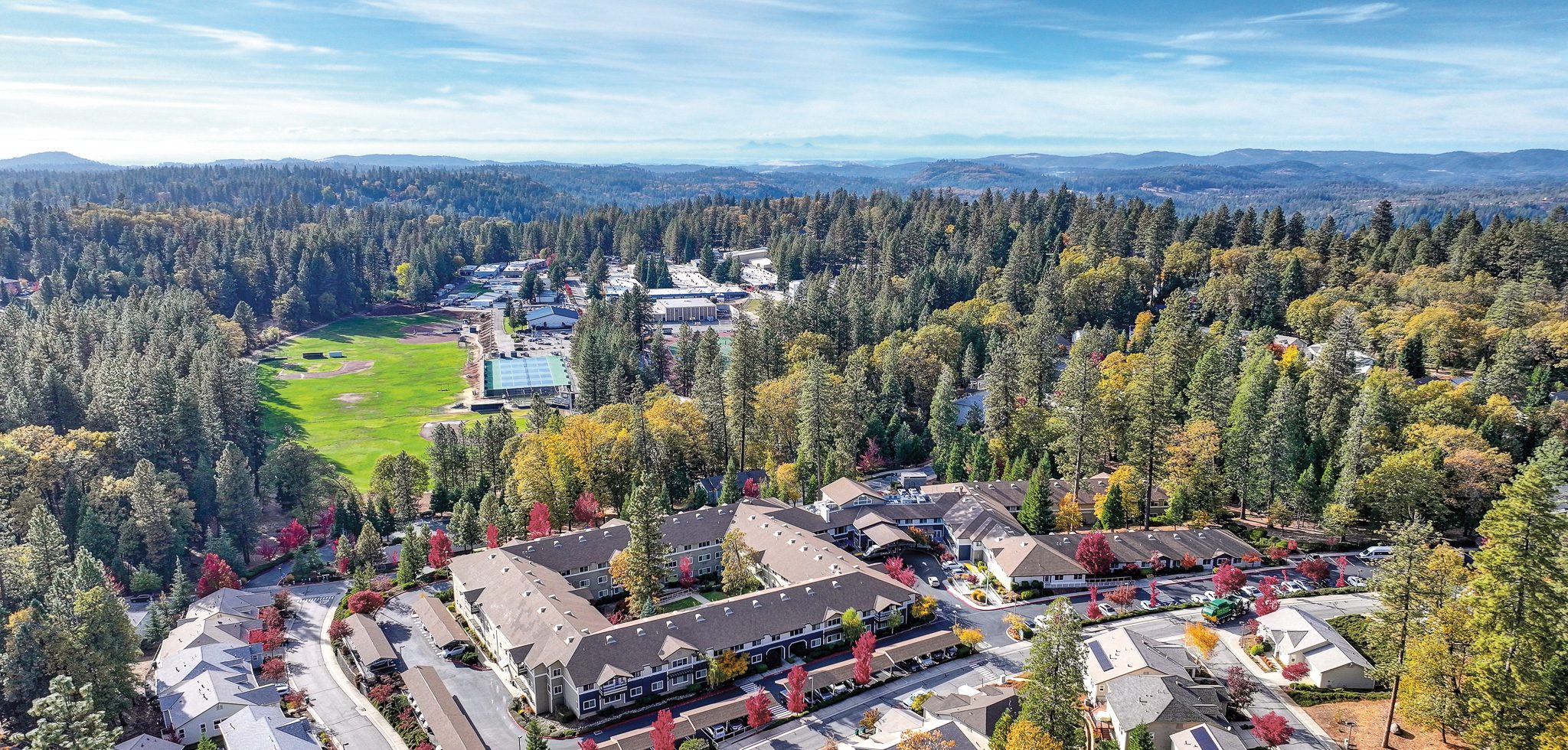 Aerial view of a residential neighborhood at About Eskaton Village Grass Valley, surrounded by dense forest, with a large green sports field and several buildings in the background under a blue sky.