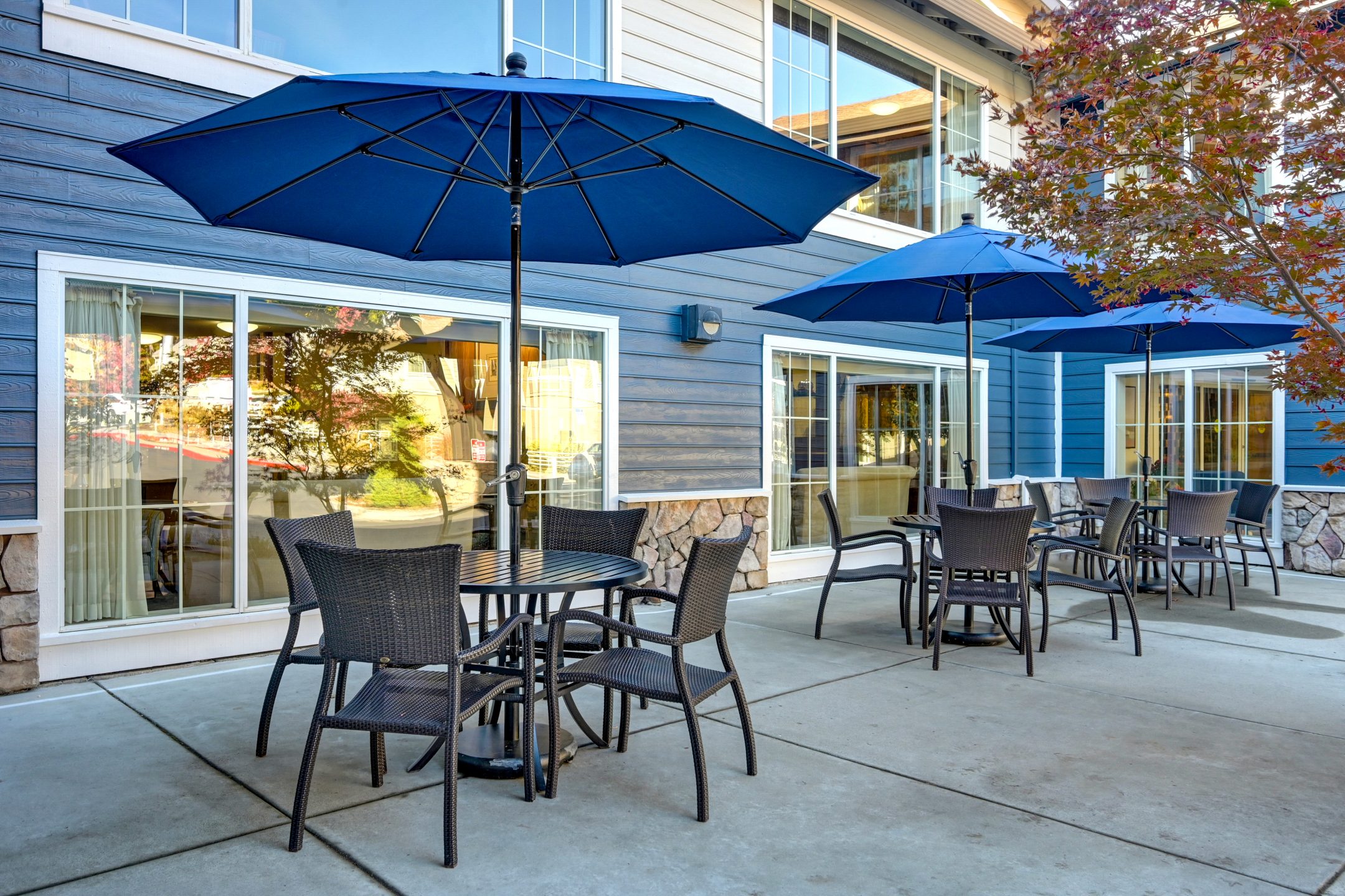 Outdoor patio area with several tables and chairs under large blue umbrellas, set on a concrete surface beside a modern building with large windows and blue siding, offering inviting amenities for relaxation or gatherings.