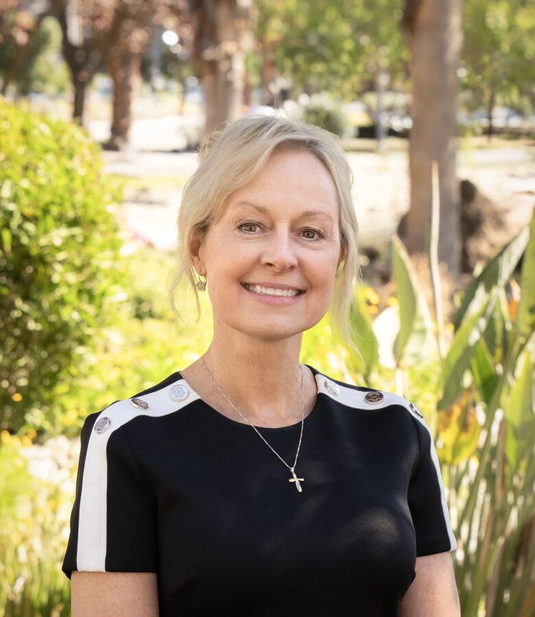 A smiling woman with blonde hair, wearing a black top with white stripes and a cross necklace, stands outdoors in the sunny, green park at Eskaton Village Grass Valley senior living, surrounded by trees and bushes.