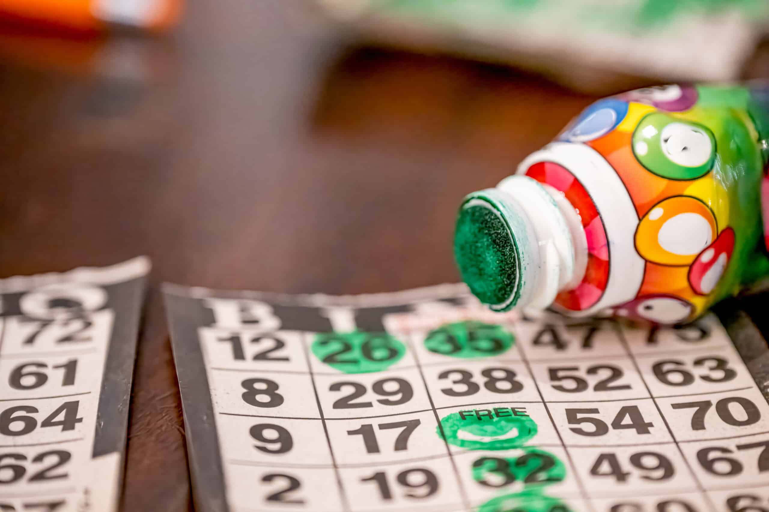 A close-up of a bingo card with several numbers marked in green ink sits ready for Designer Bag Bingo. A colorful bingo dauber, its tip stained green, rests on the table beside the card, while another bingo card is partially visible.