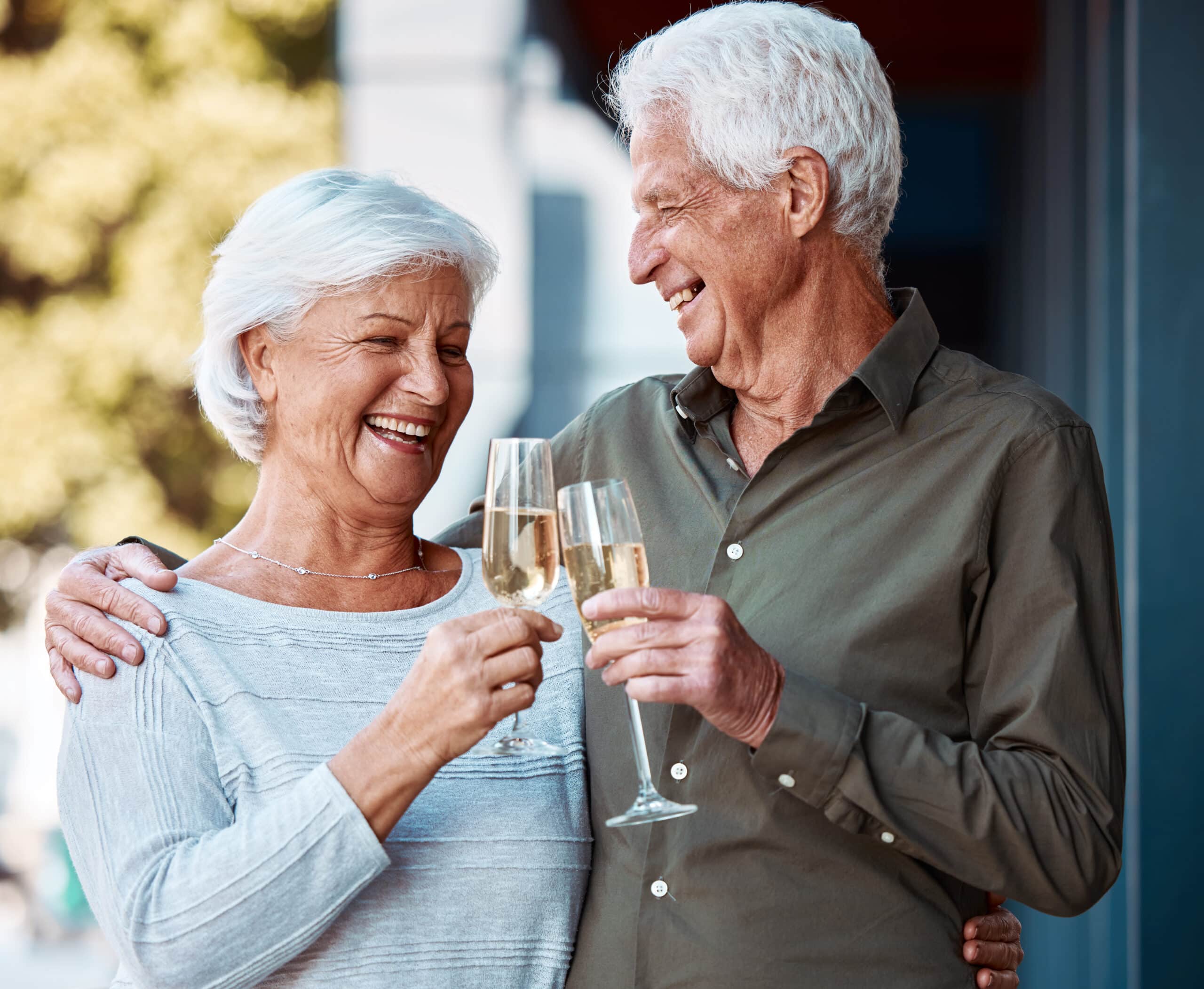An elderly couple smiles and laughs while clinking champagne glasses outdoors, enjoying snacks and each other's company. The man has his arm around the woman, and both appear happy and relaxed.