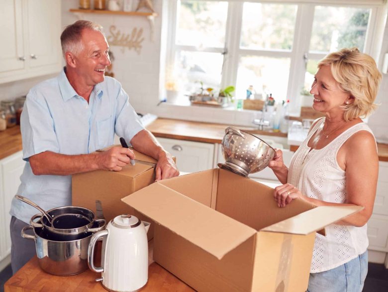Smiling older couple in a bright kitchen packing dishes into cardboard boxes, preparing to move or organize for independent living.