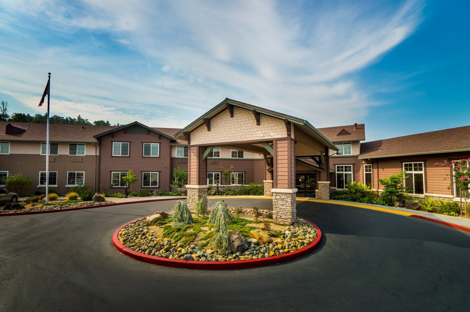 A large, modern building with brown siding and a covered entrance, surrounded by landscaped grounds and a circular driveway with a rock garden and small bushes under a partly cloudy sky.