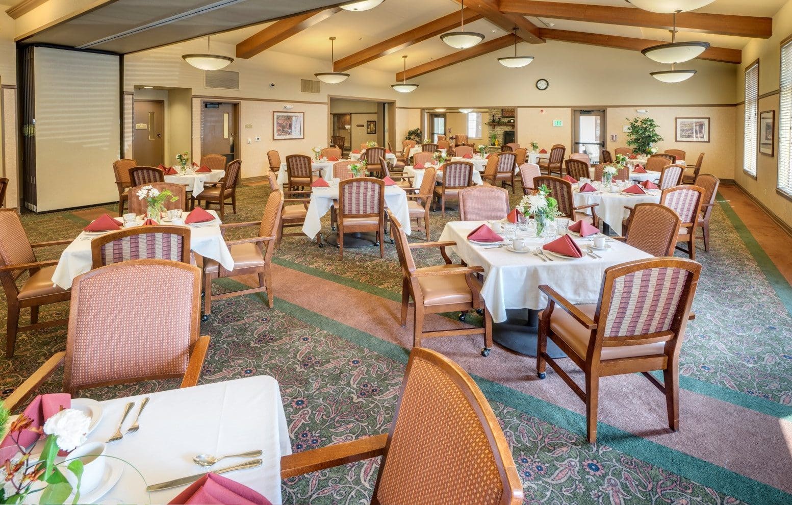 A spacious dining room set for a memorable culinary experience, featuring tables with white cloths, red napkins, floral centerpieces, and wooden chairs. High ceilings with exposed beams and large windows complete the inviting atmosphere.