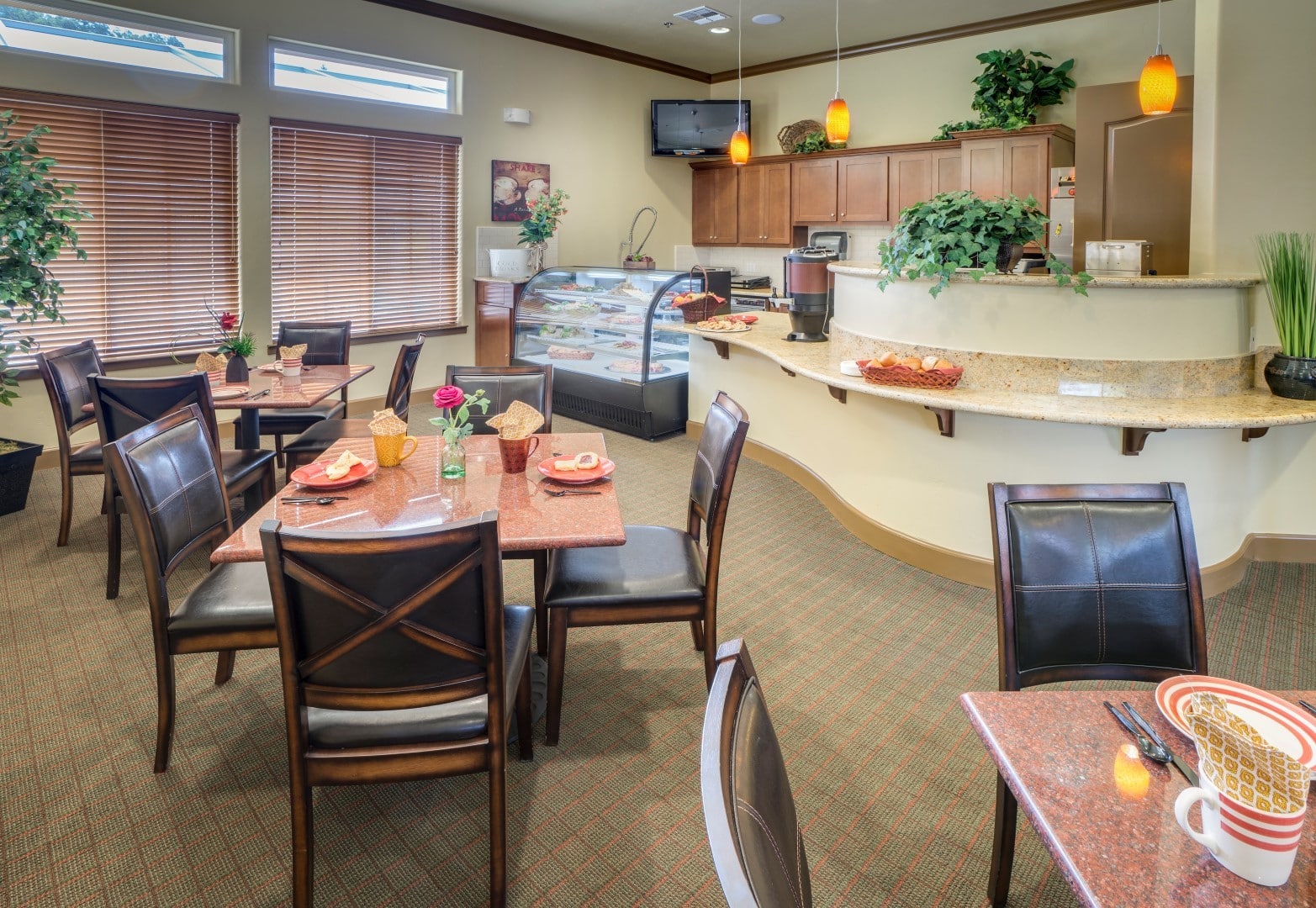 A cozy café with tables set for dining, dark wooden chairs, and a counter with pastries. Warm lighting, large windows with blinds, green plants, and decorative accents offer inviting living options in a welcoming atmosphere.