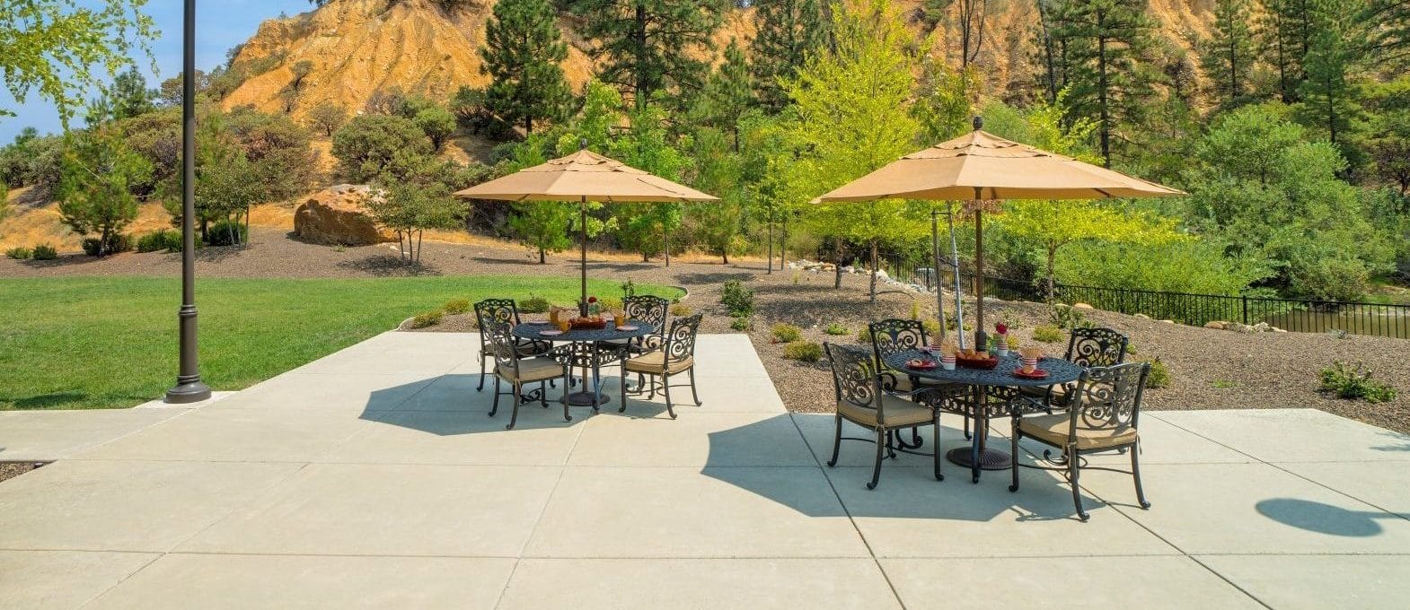 Patio with metal tables and chairs under beige umbrellas, set for a delightful culinary experience, overlooks a grassy yard and rocky, tree-covered hillside under a blue sky with scattered clouds.