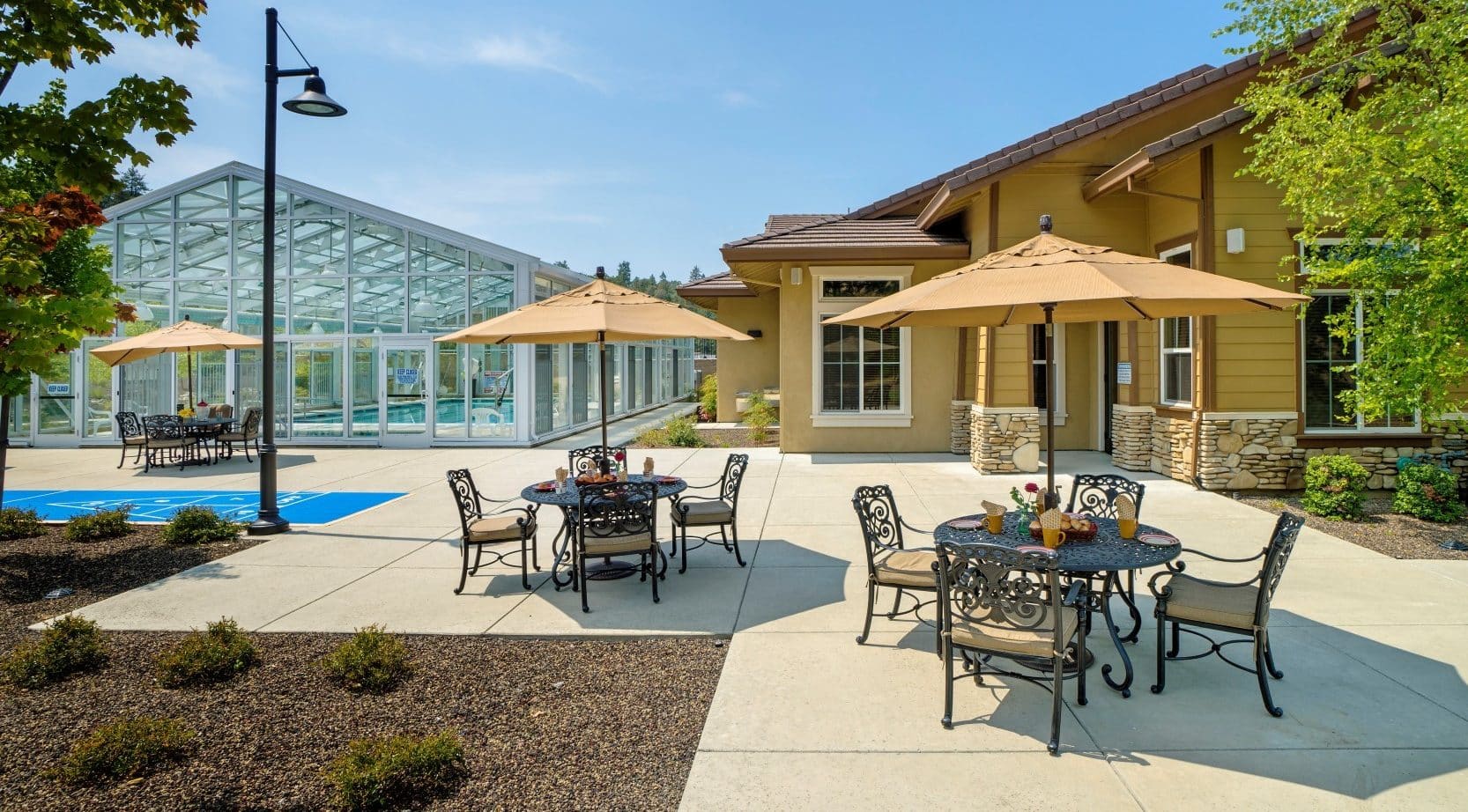 Outdoor patio with metal tables and chairs under tan umbrellas next to a tan building and glass greenhouse. This inviting assisted living area is beautifully landscaped with trees and shrubs under a clear blue sky.