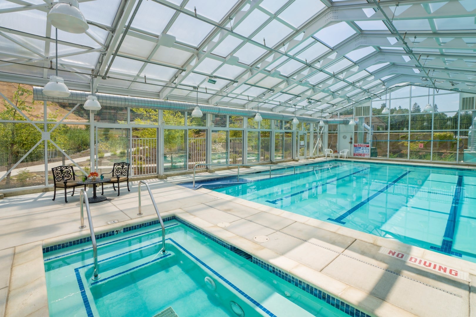 Indoor pool area in the assisted living community boasts large windows and a glass ceiling, a swimming pool, hot tub, poolside table with two chairs, and scenic views of trees and hills outside. No Diving is clearly marked on the poolside.