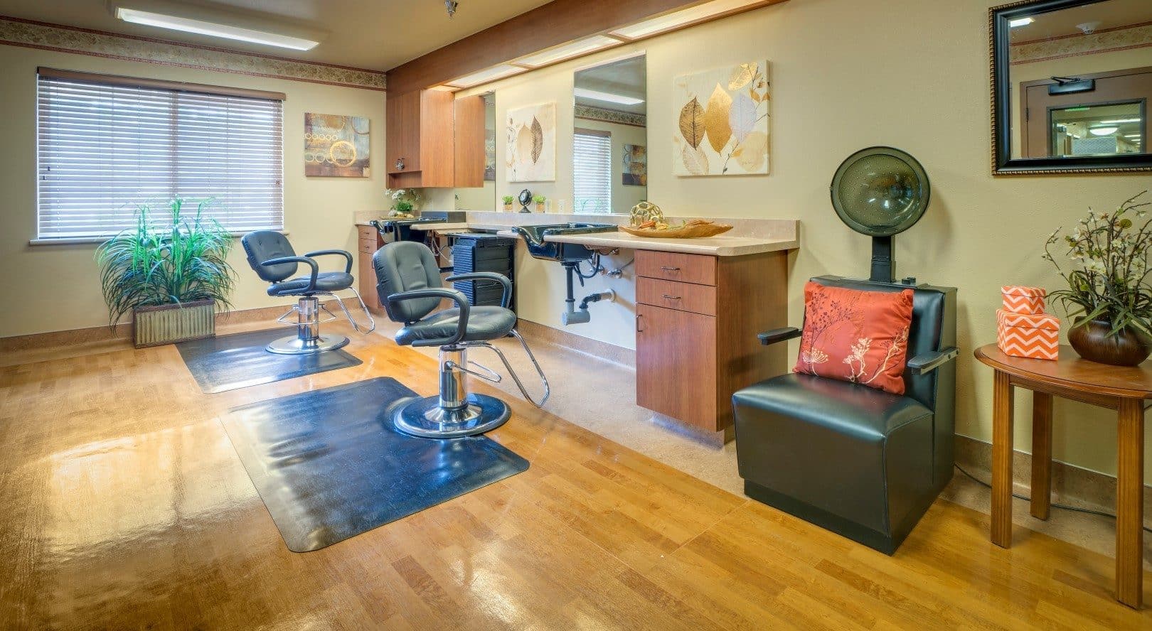A well-lit hair salon with two styling chairs, a counter with mirrors, and a plant by the window offers modern decor and wooden floors—an inviting space that could inspire your living options. A dryer chair with a red cushion completes the look.