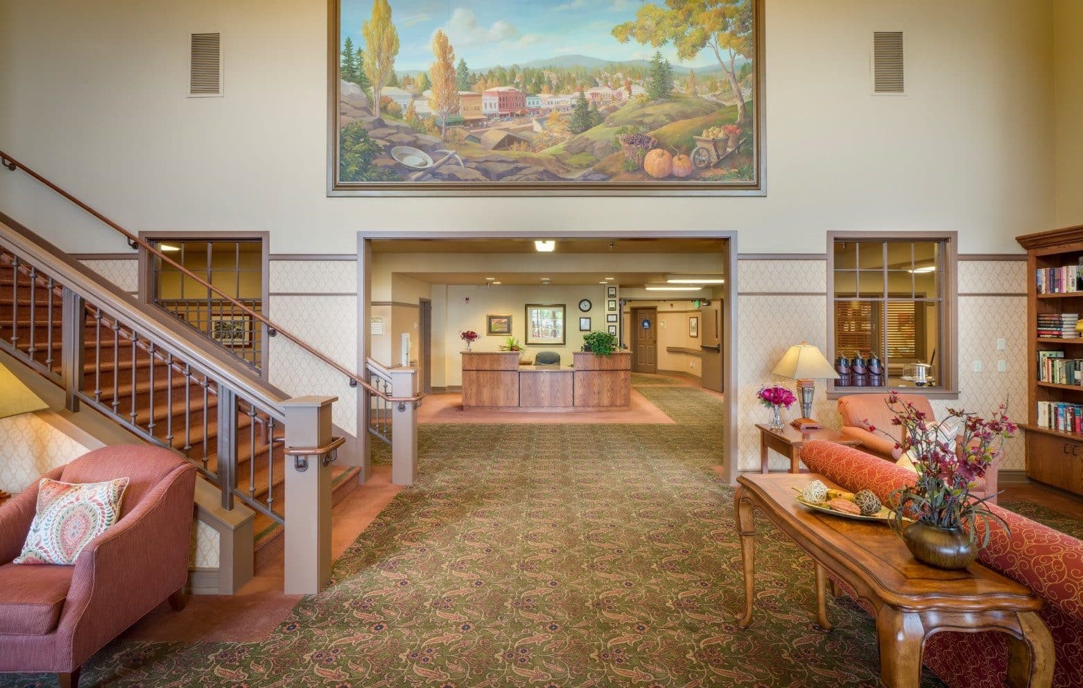 A spacious, warmly lit lobby designed for assisted living features a large landscape painting above a wooden reception desk, with cozy seating, patterned carpet, bookshelves, and a staircase on the left. Decorative flowers and lamps add a welcoming touch.