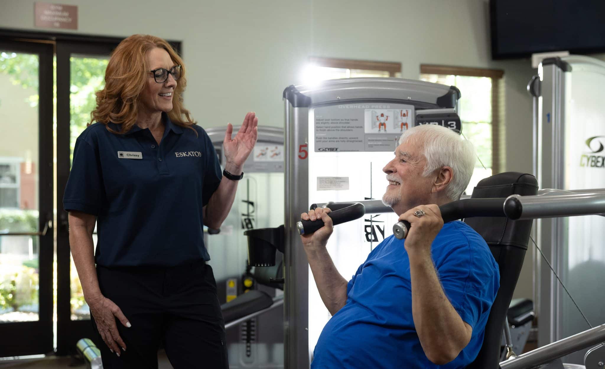 A smiling older man lifts weights on a gym machine while a woman in a staff uniform stands nearby, talking to him and gesturing with her hand. They are in a bright, modern fitness center.